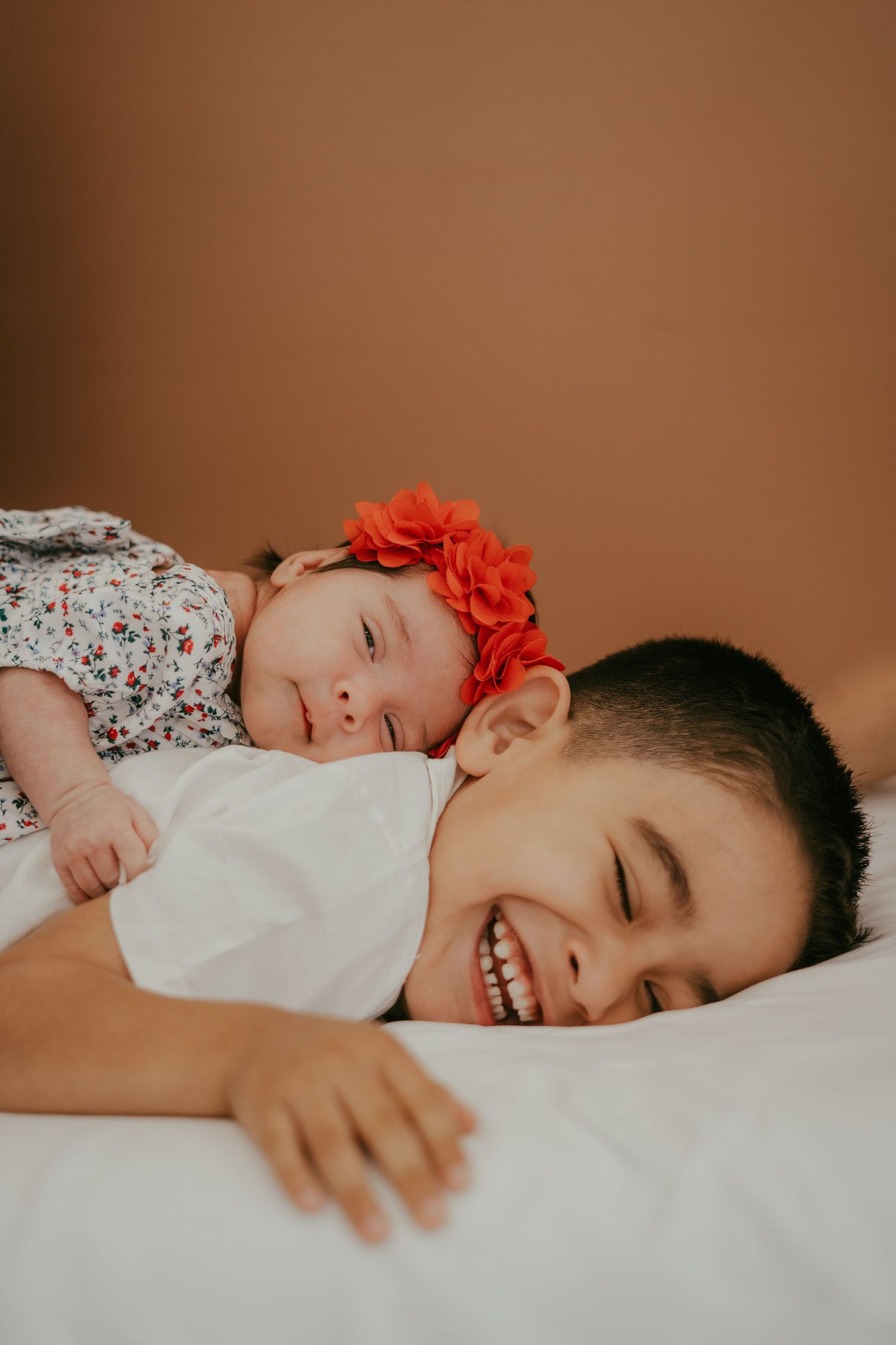 Joyful older sibling laughs with eyes closed as newborn baby girl with orange floral headband smiles on white bedding.