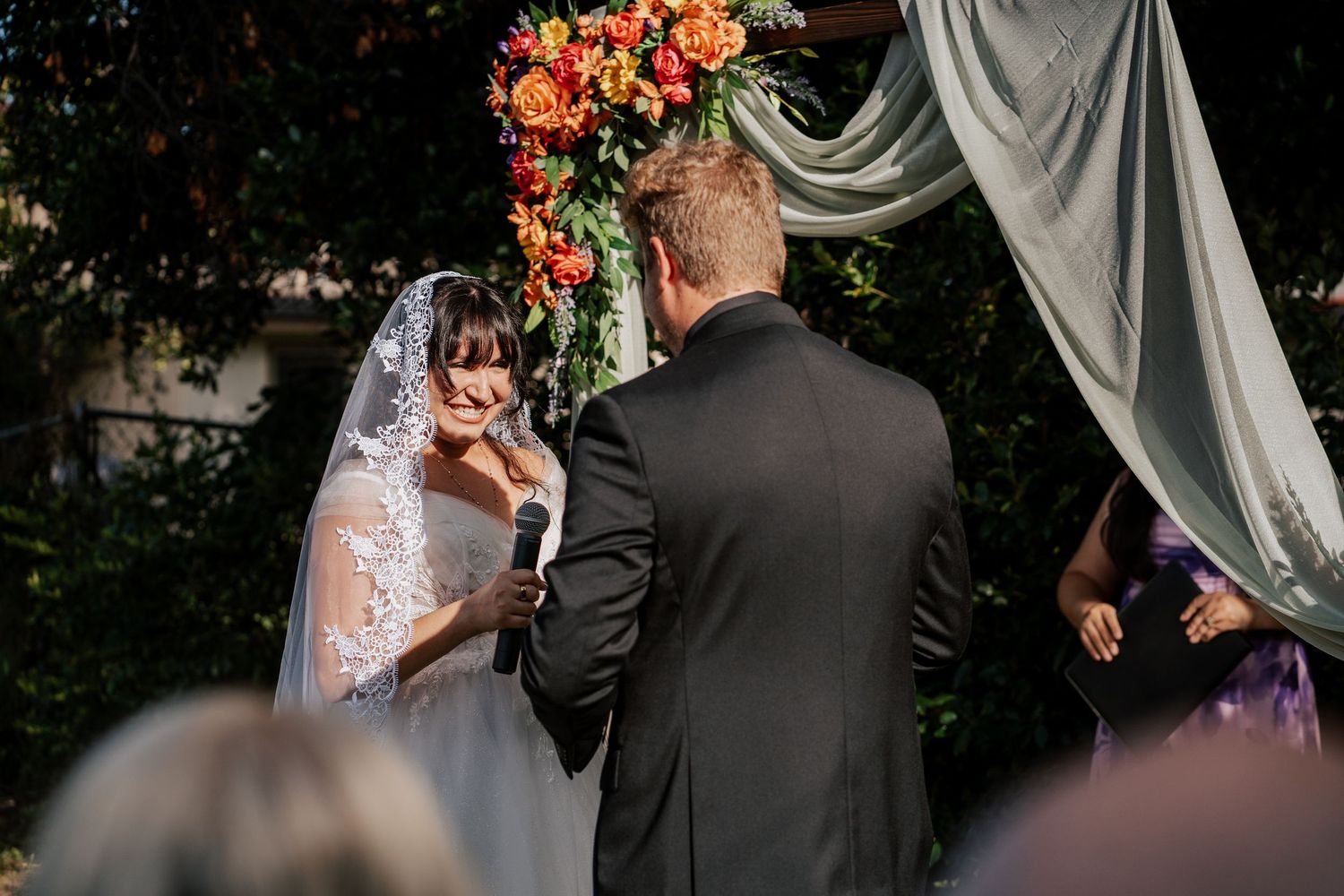 Bride reading vows to groom at outdoor wedding ceremony under floral arch with orange and red blooms.