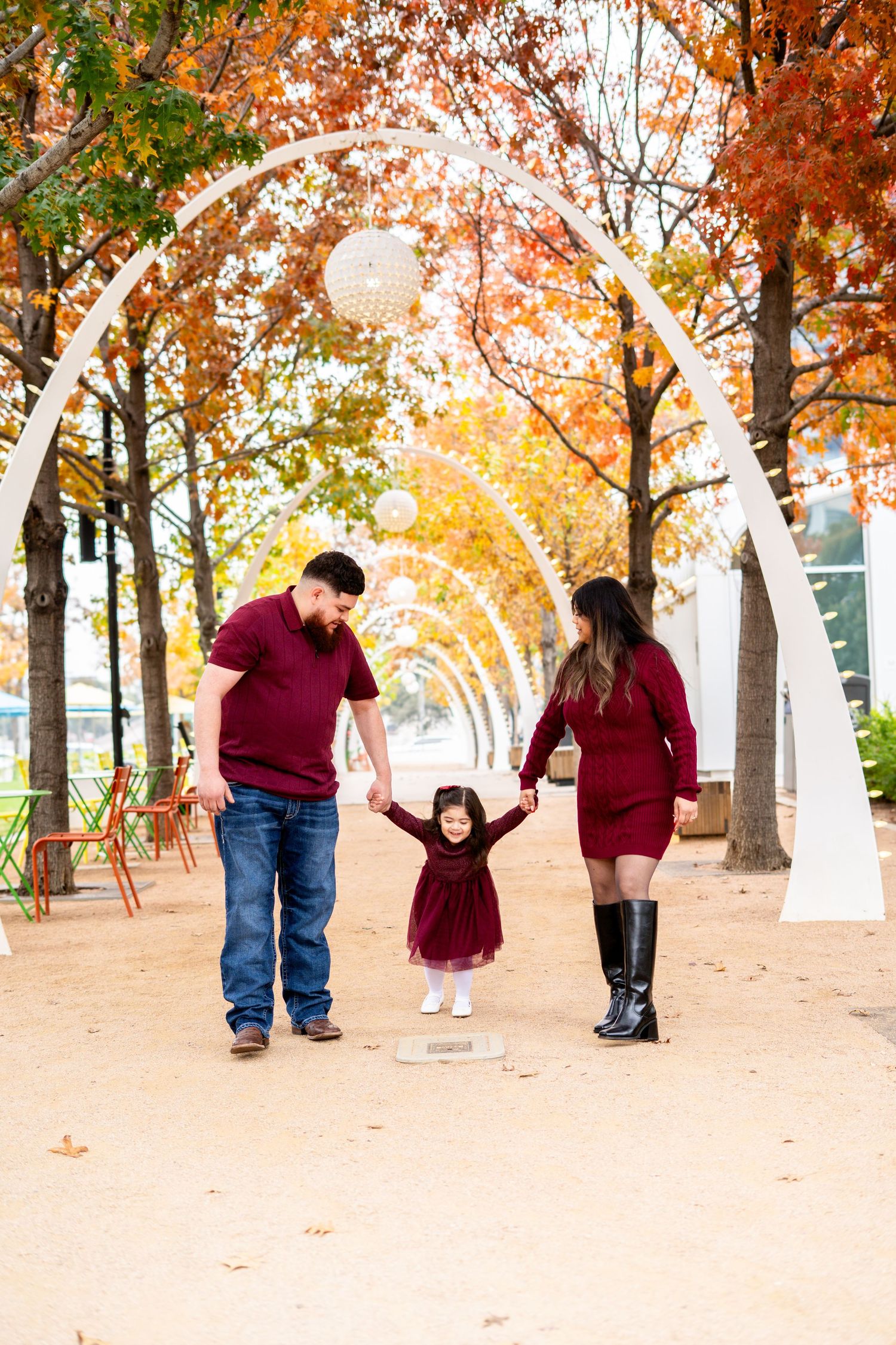 Family of three in burgundy outfits holding hands under white arches with fall foliage in a park.