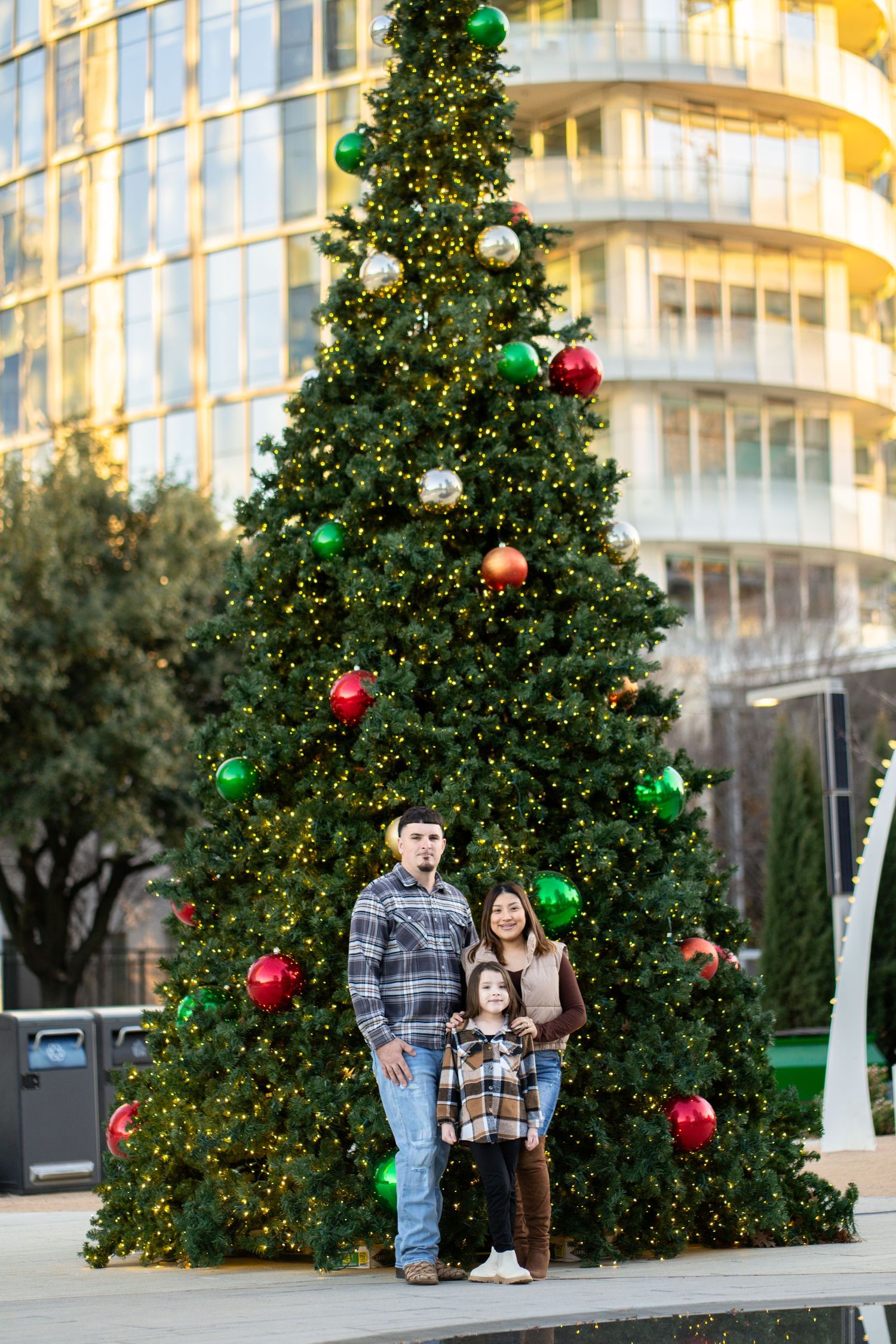 Family of three stands together in front of a tall outdoor Christmas tree decorated with colorful ornaments.