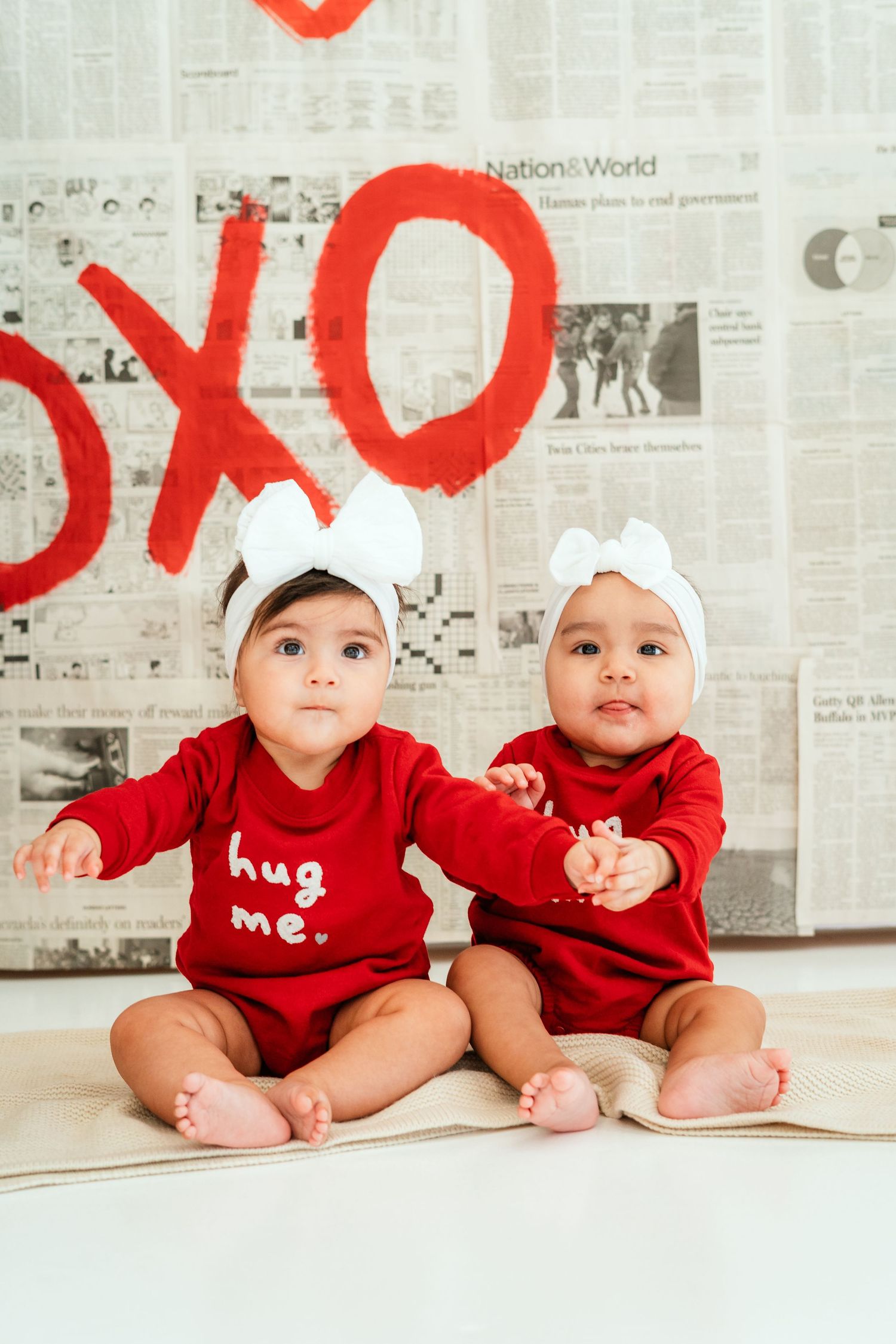 Two babies in red 'hug me' onesies sit before Valentine's XOXO backdrop.