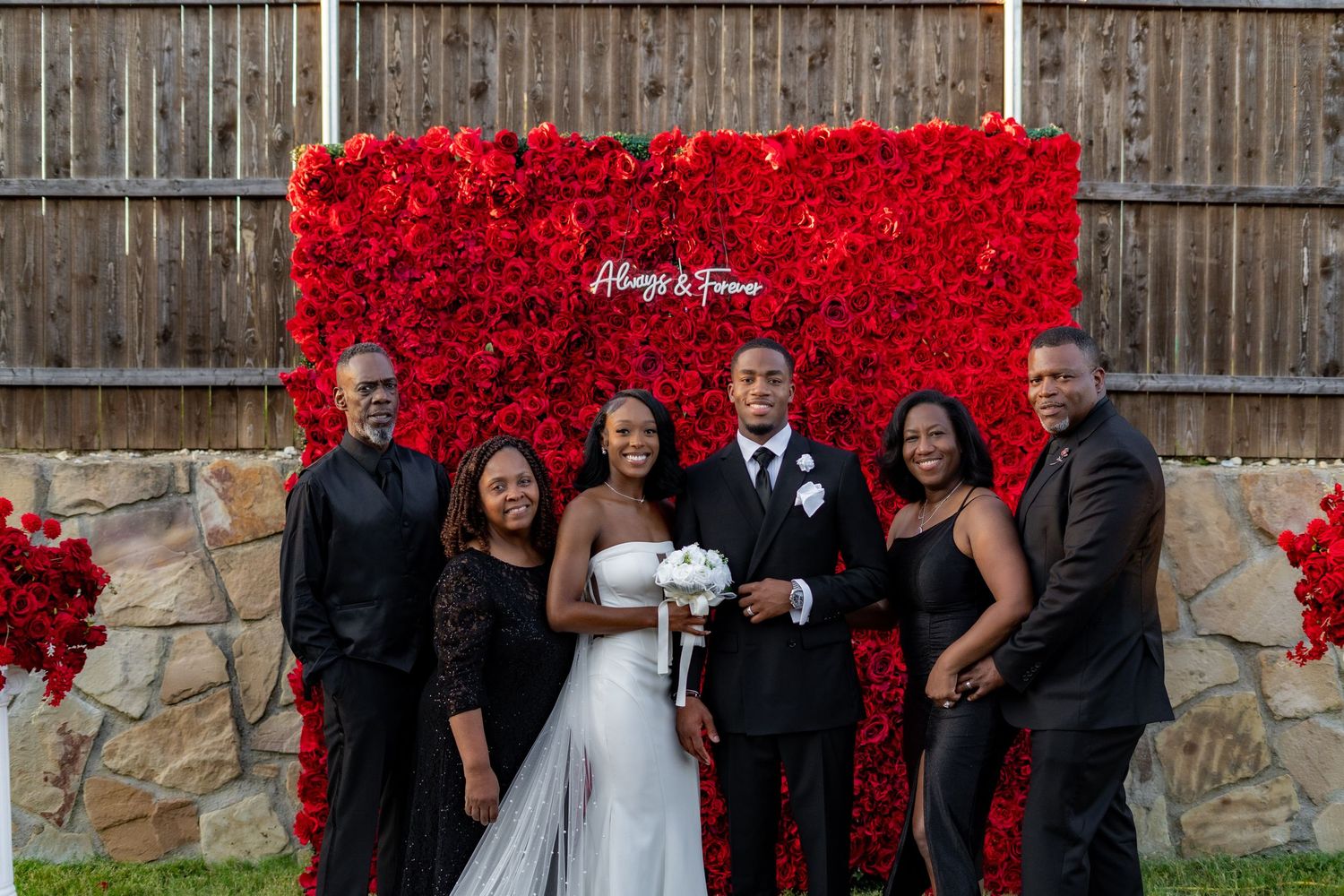 Bride and groom pose with four family members before an Always & Forever red rose flower wall.