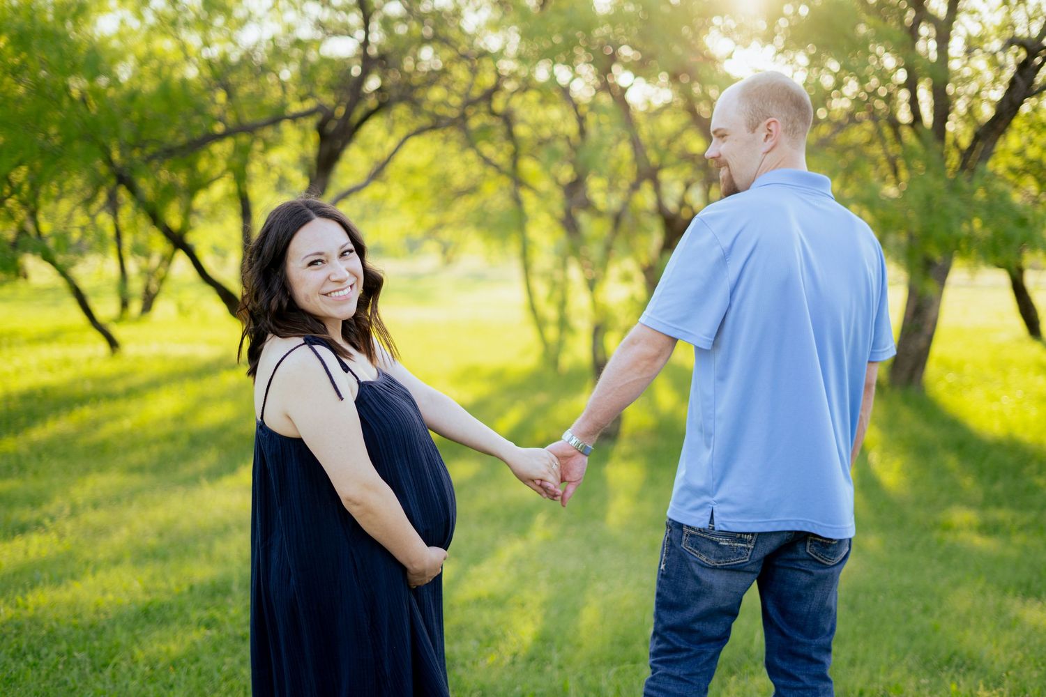 Pregnant woman in navy dress holding hands with partner while walking in a sunny park.