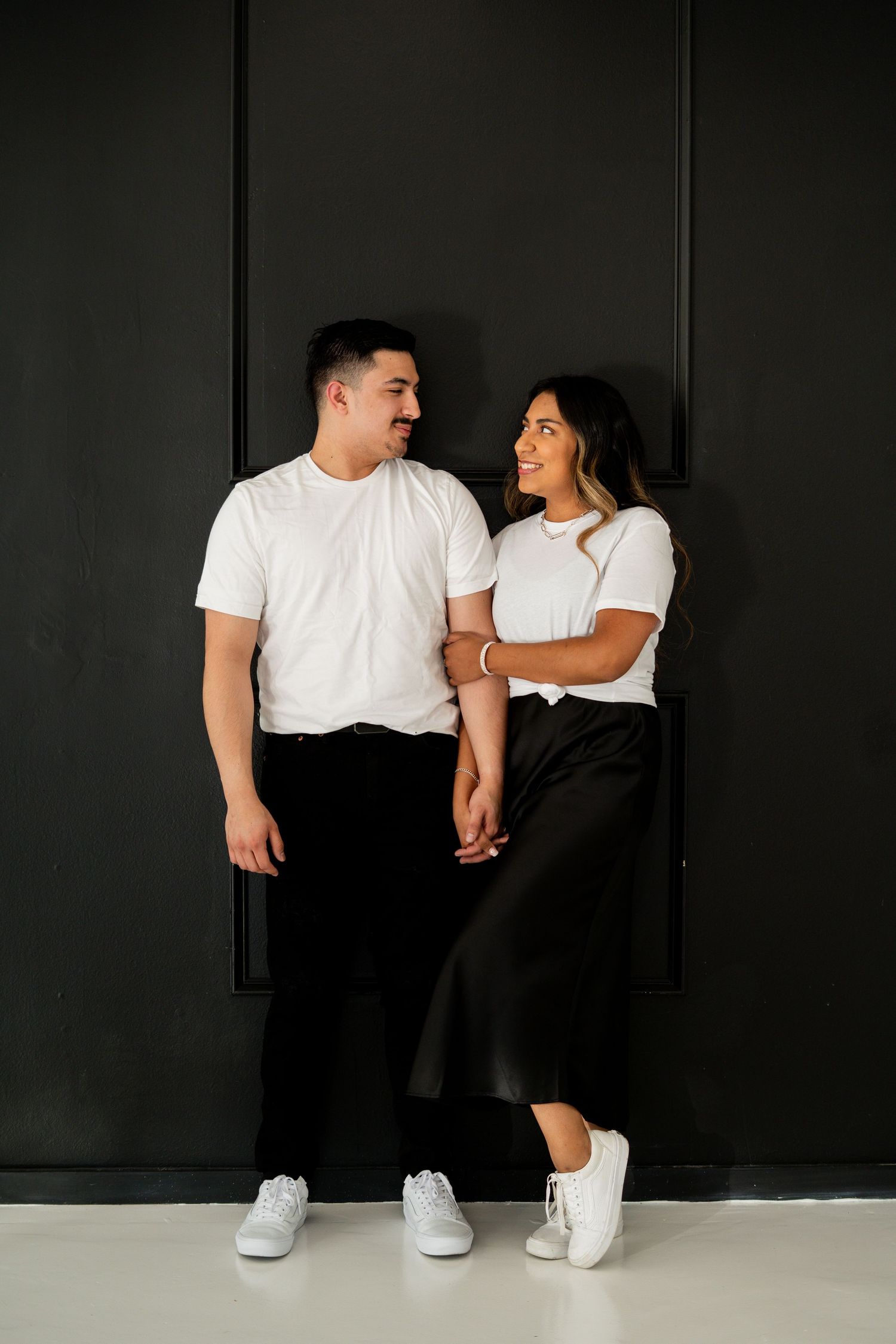 Smiling couple in matching white tees and black bottoms holding hands against a dark wall.