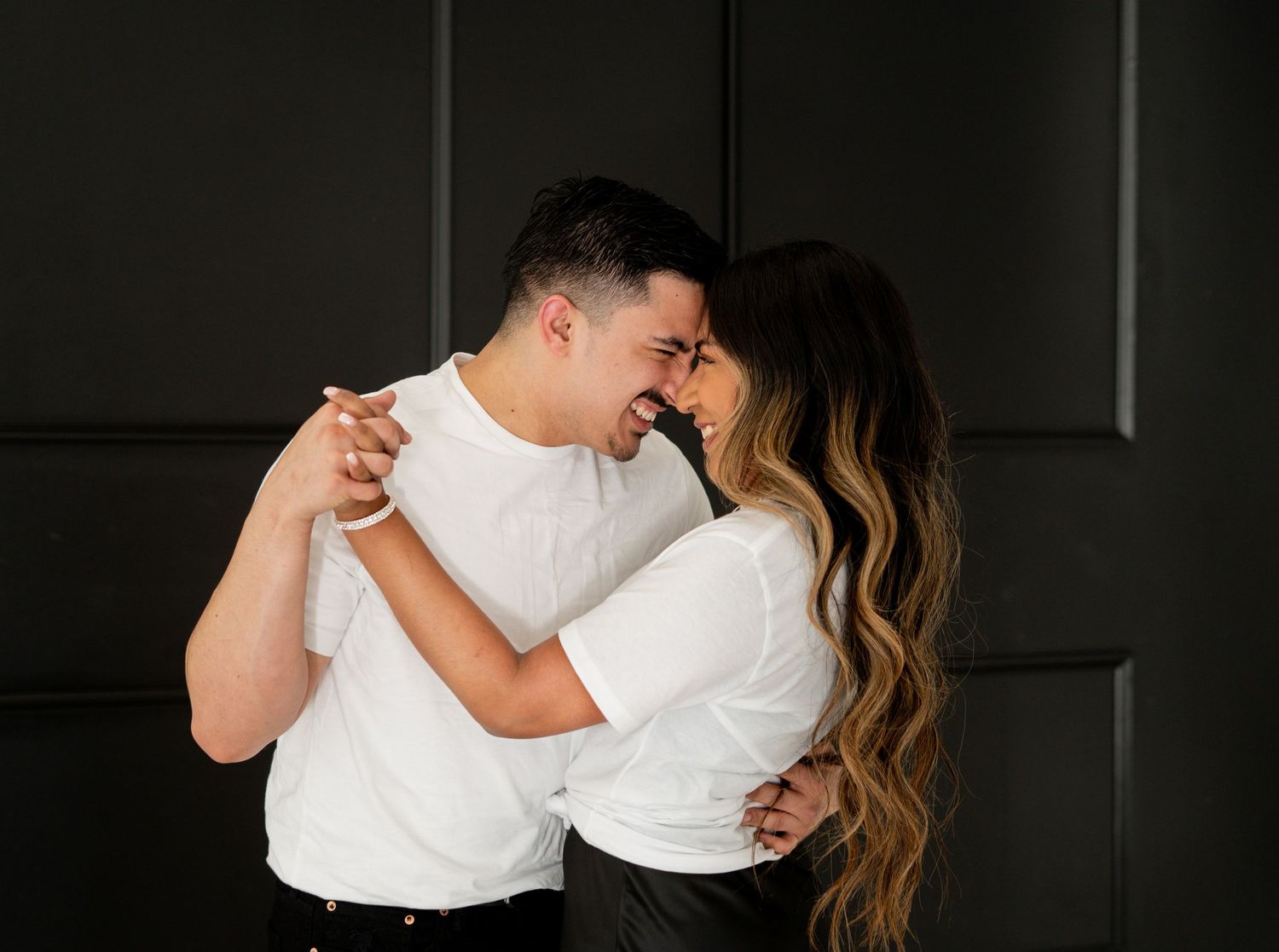 Couple in white shirts touching foreheads and embracing against a dark wall.