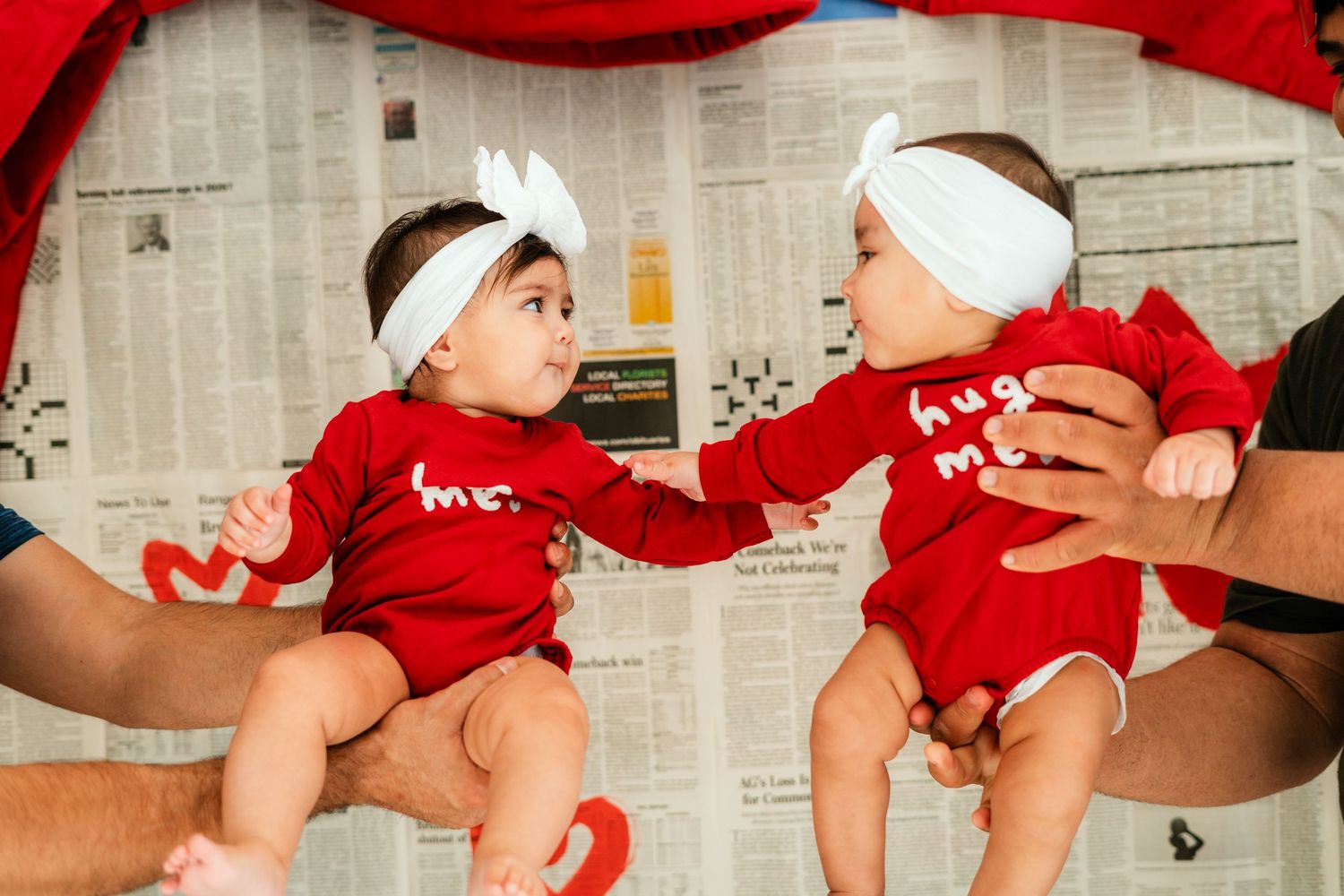 Two babies in red 'hug me' onesies and white bows face each other.