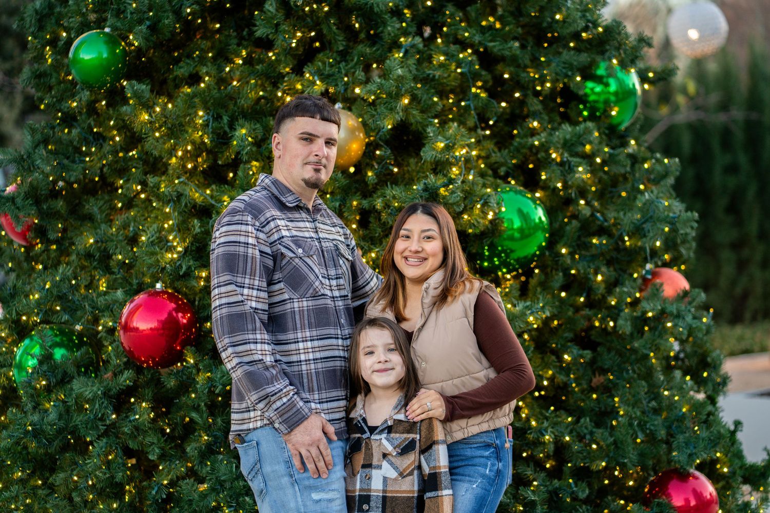 Family of three poses in front of a large decorated Christmas tree with colorful ornaments outdoors.