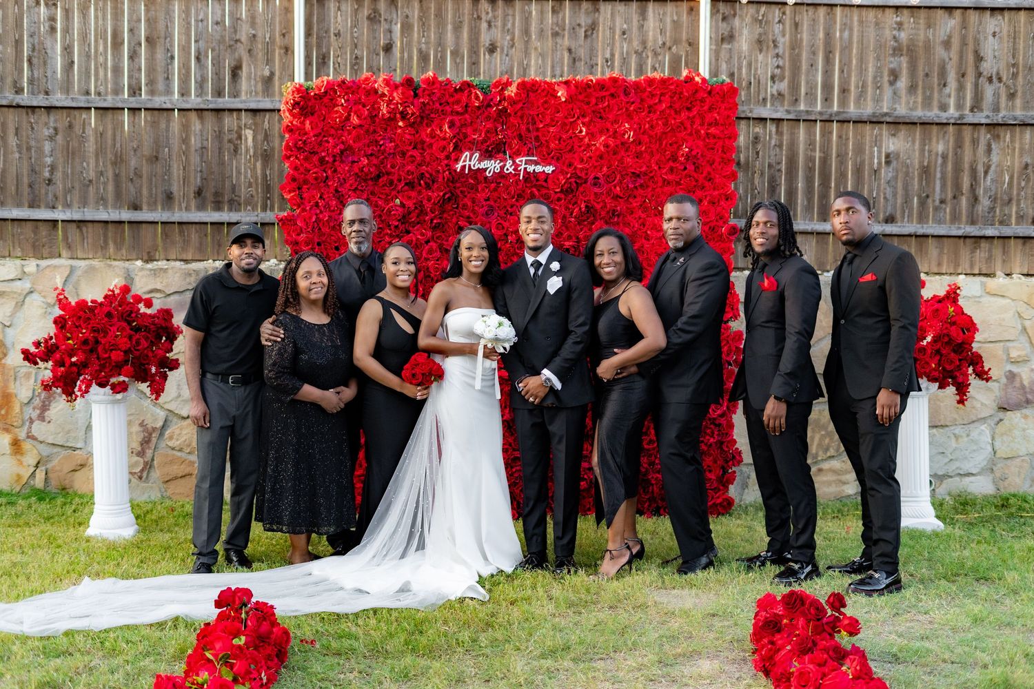 Wedding party of ten poses in front of red rose backdrop with floral arrangements on white pillars.