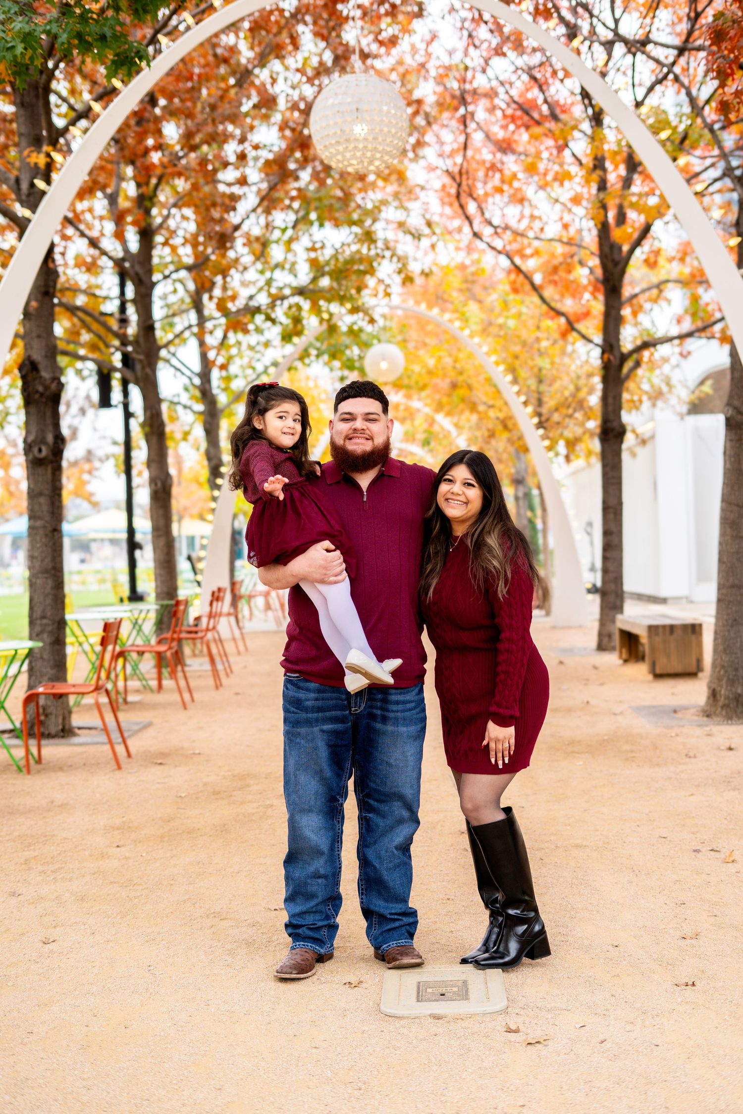 Family of three in matching burgundy outfits posing under decorative arches with colorful fall trees.