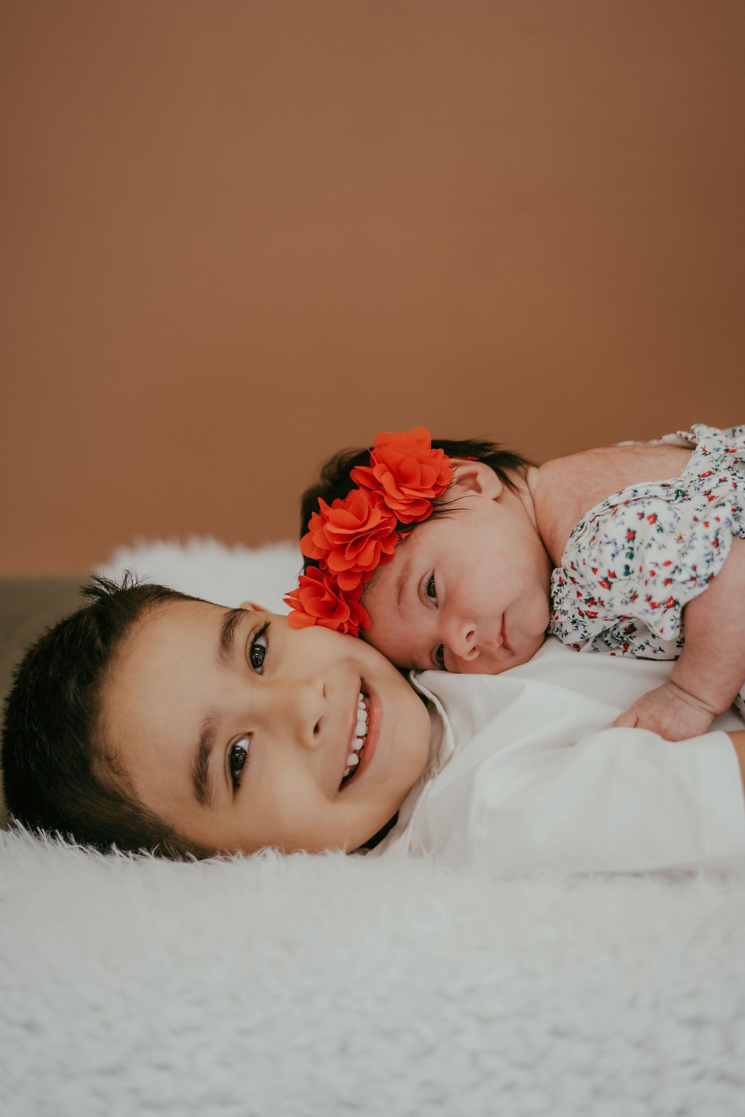 Older child smiles at camera while newborn baby in floral outfit and red flower headband rests beside them on white fur.