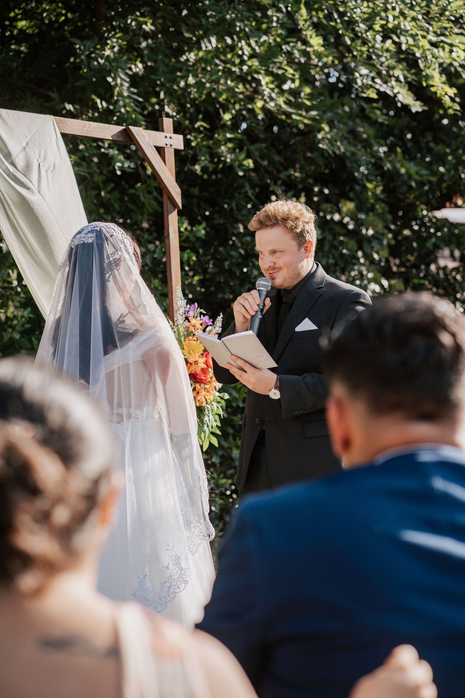 Groom reads vows at outdoor wedding ceremony with bride in lace veil before wooden arch with flowers.
