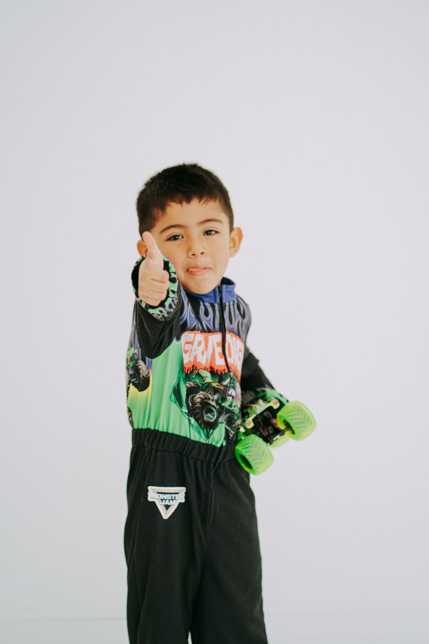 Young boy in Monster Jam racing costume gives thumbs up while holding green toy monster truck.