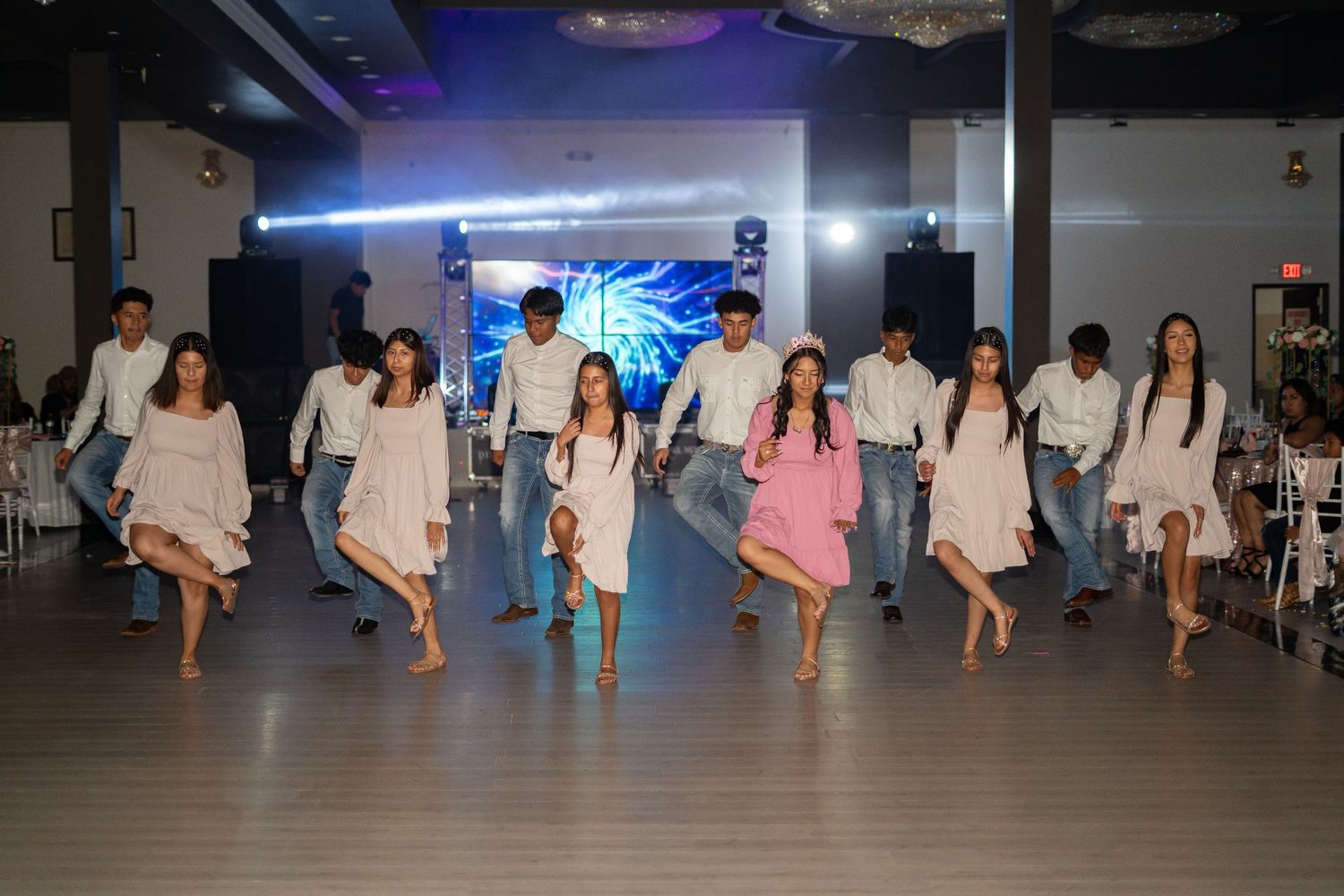 Quinceañera court performs choreographed dance in white outfits with girl in pink dress at center.