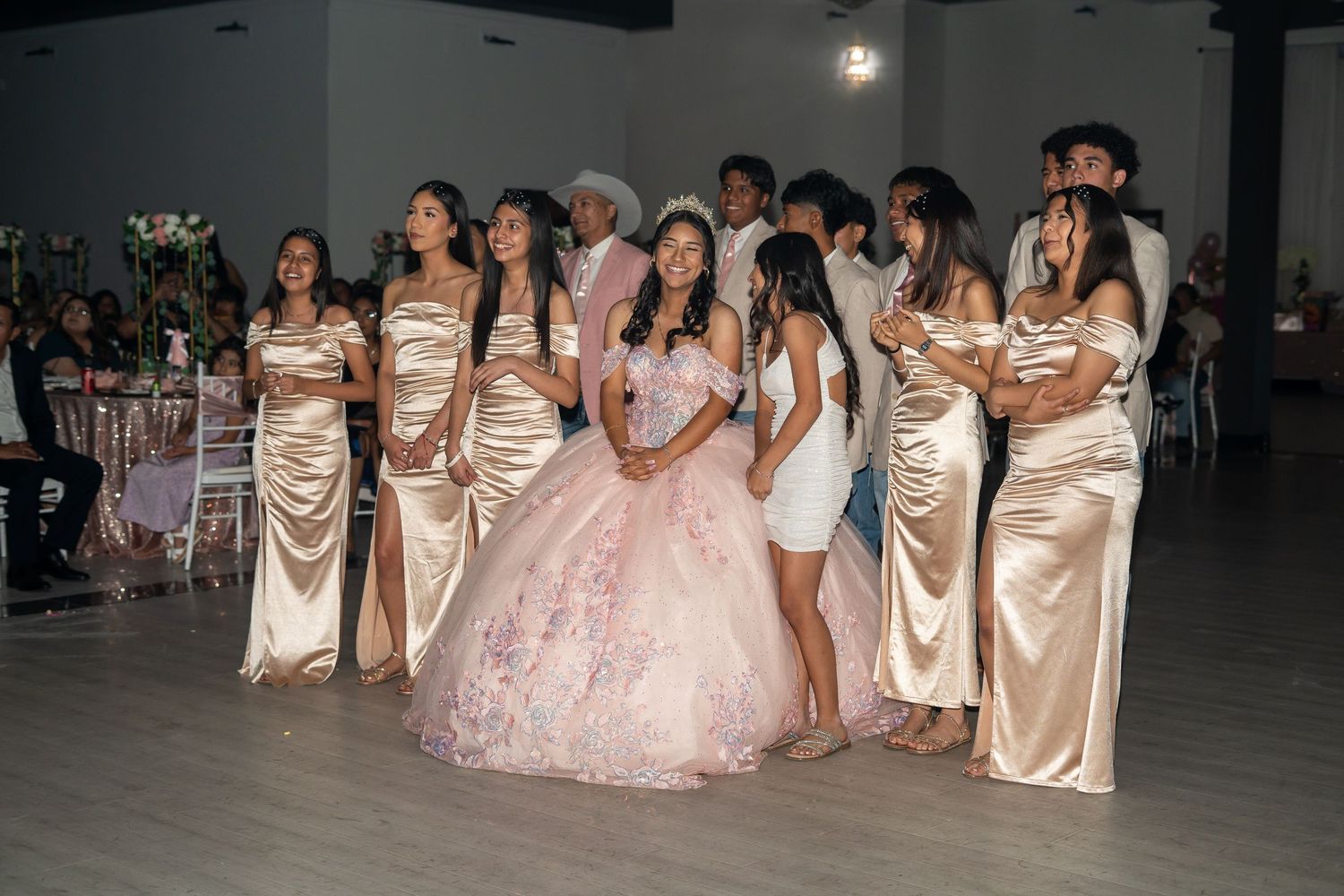 Quinceañera girl in pink gown poses with chambelanas in gold satin dresses on decorated dance floor.