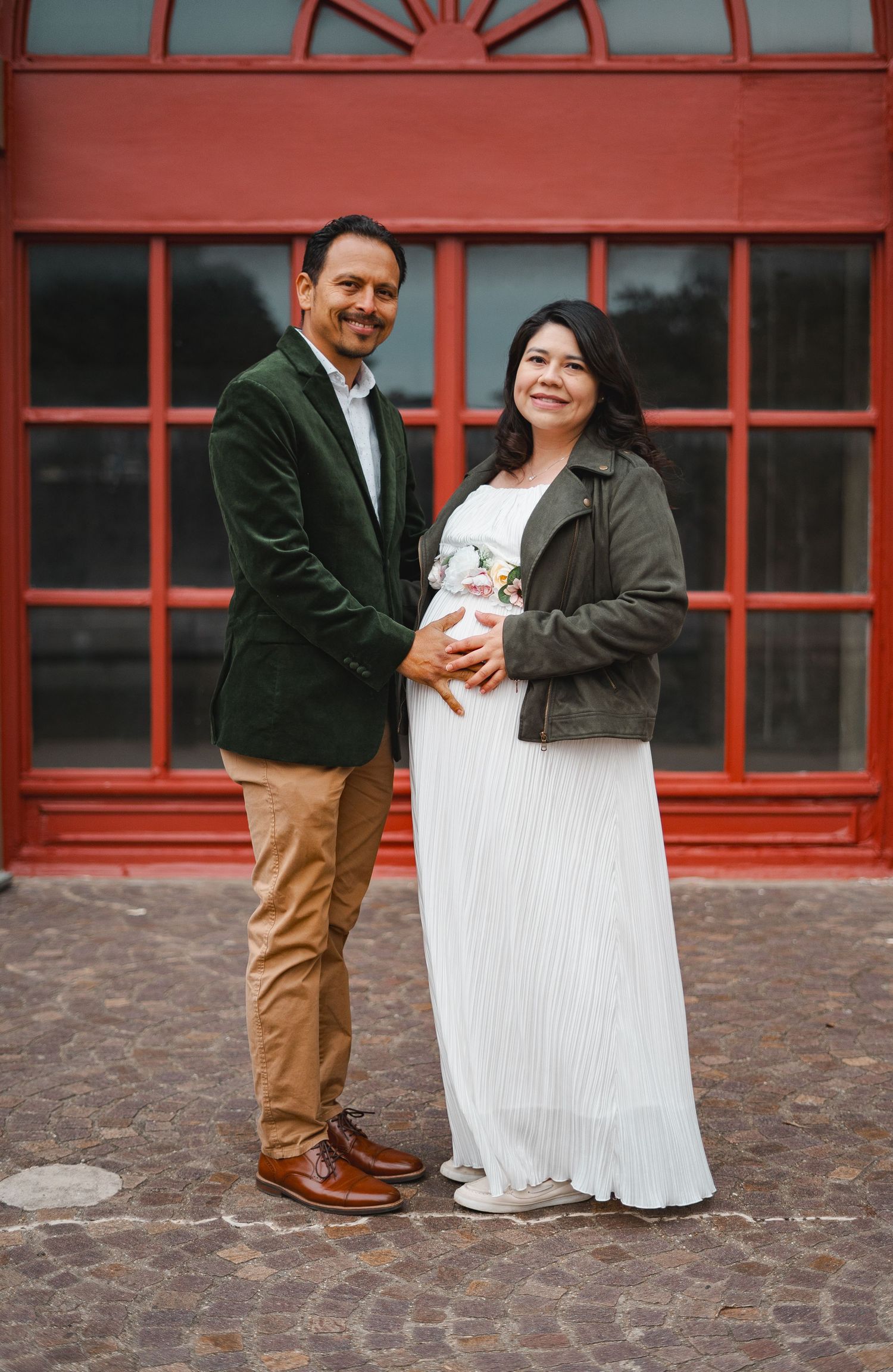 Happy couple posing for maternity photos in front of red building, woman in white dress.