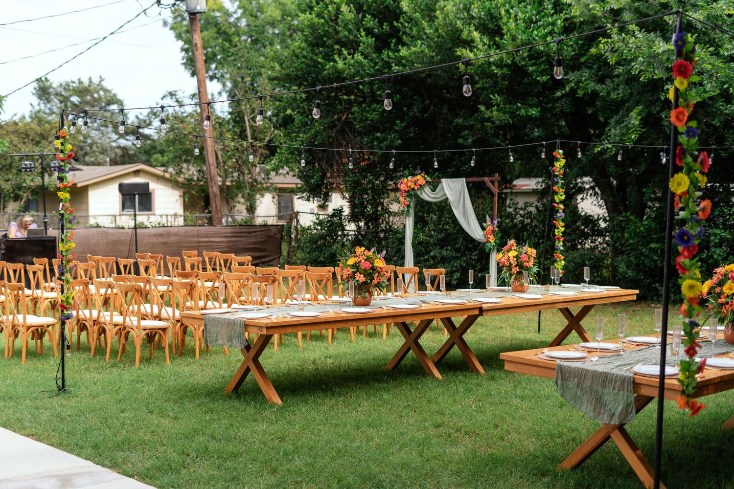 Outdoor wedding reception setup with wooden farm tables, cross-back chairs, and floral arrangements on a green lawn.