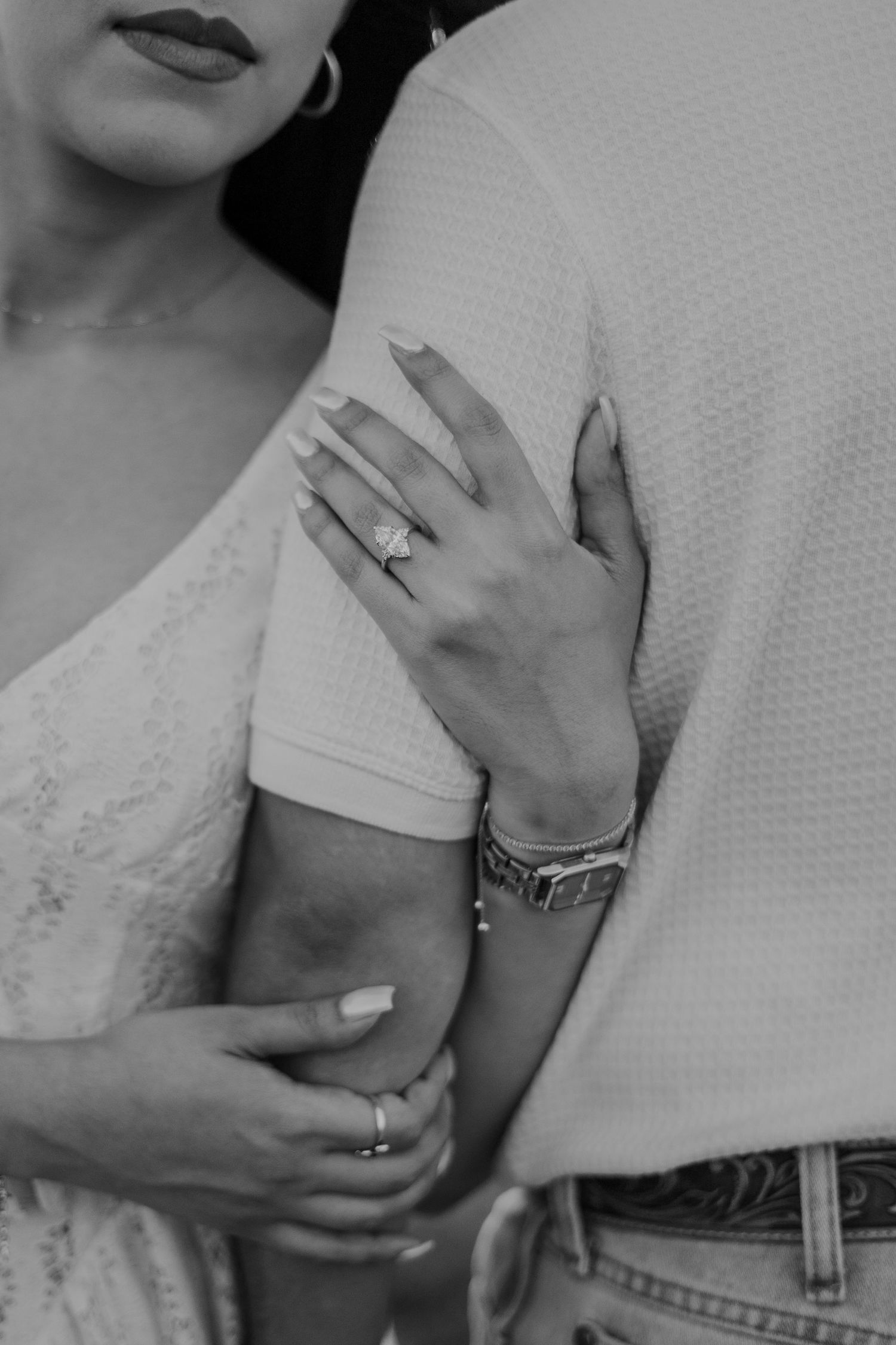 Black and white photo of couple embracing, woman's hand showing elegant engagement ring and bracelet.