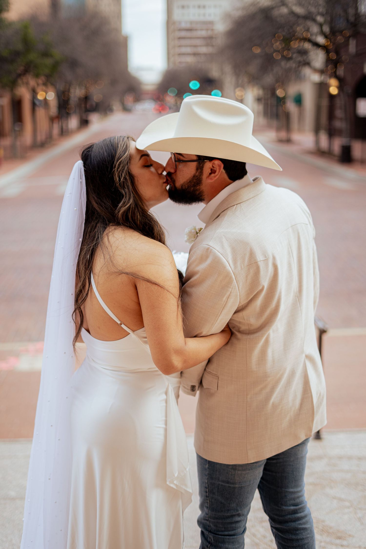 A bride in a white dress and veil kisses her groom in a cowboy hat on a city street.