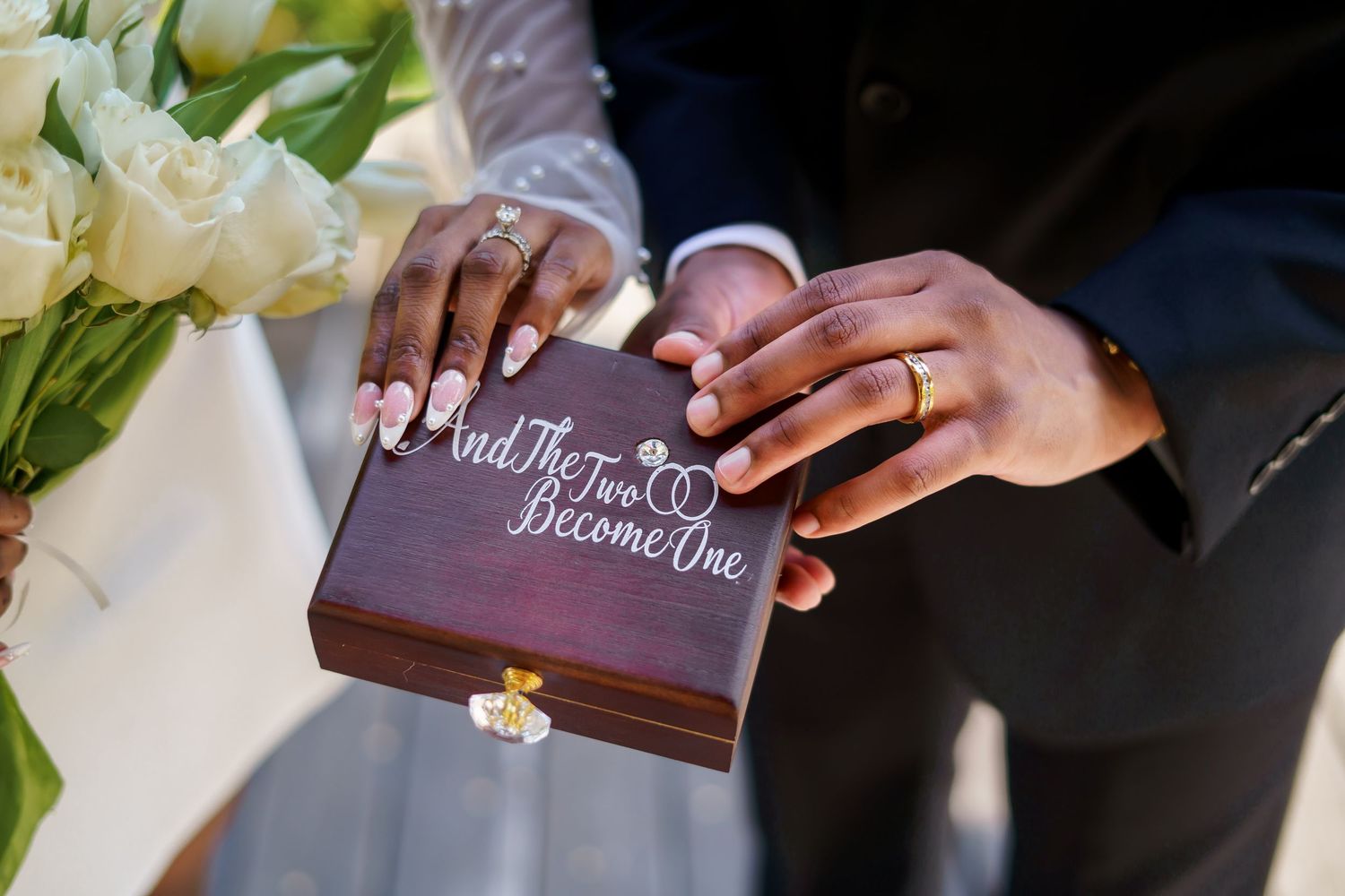 Close-up of newlyweds hands on a wooden ring box engraved with And The Two Become One.