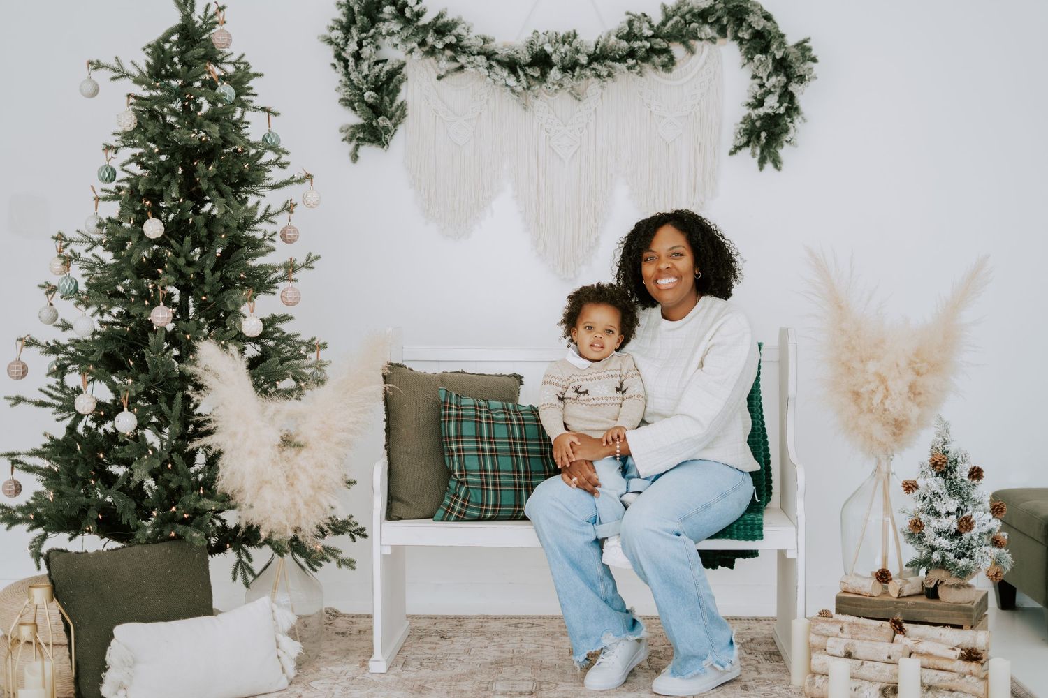 A smiling mother and toddler sit together on a white bench beside a decorated Christmas tree.
