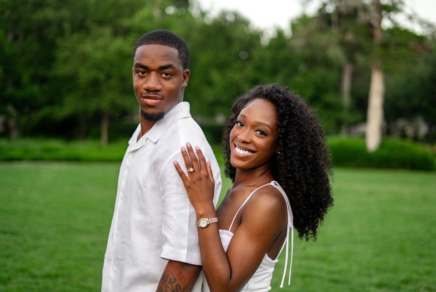 A smiling Black couple poses together outdoors, with the woman showing off her engagement ring on her hand.