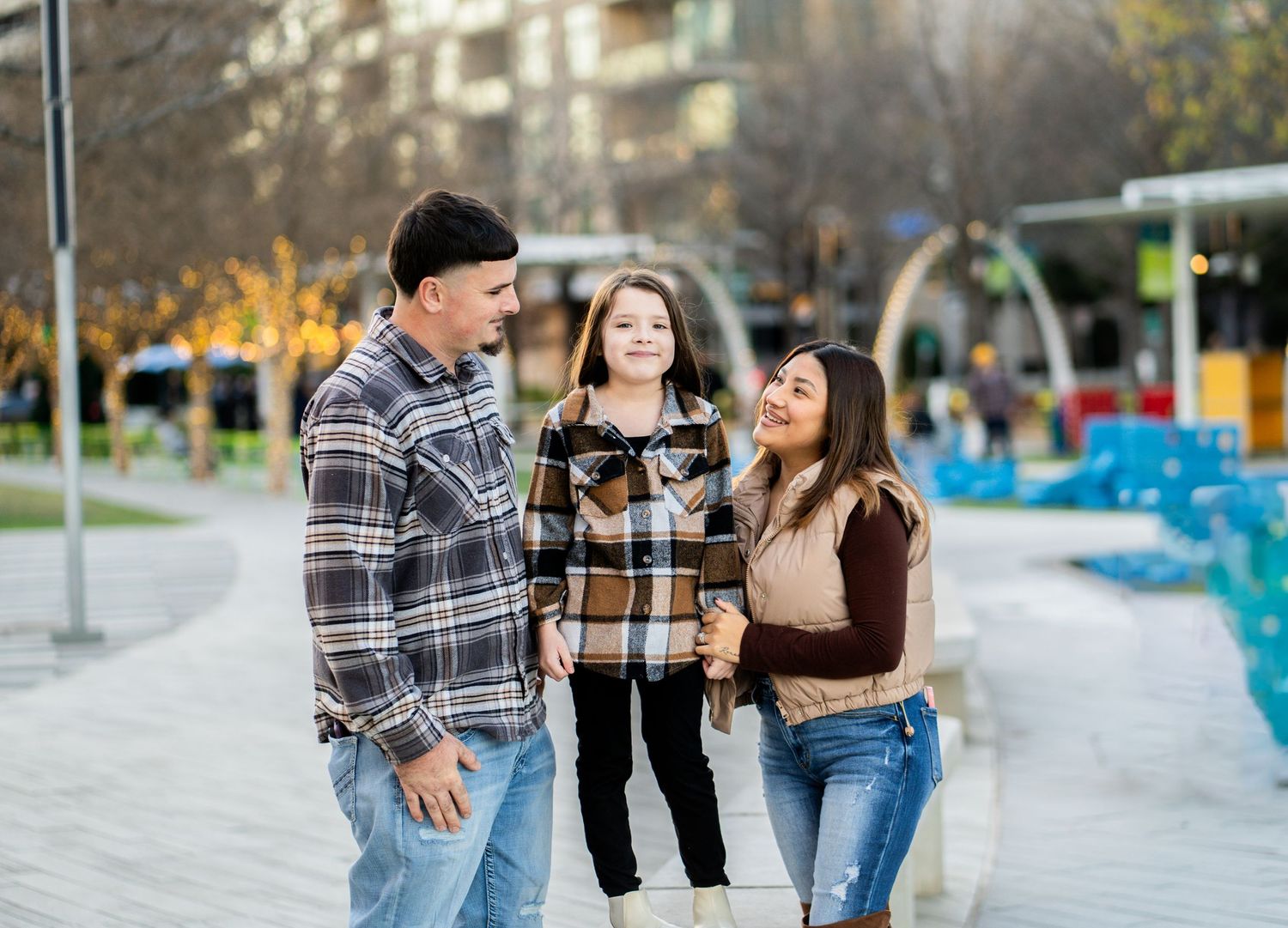 A smiling family of three enjoys a cheerful moment together on a sunny outdoor plaza walkway.