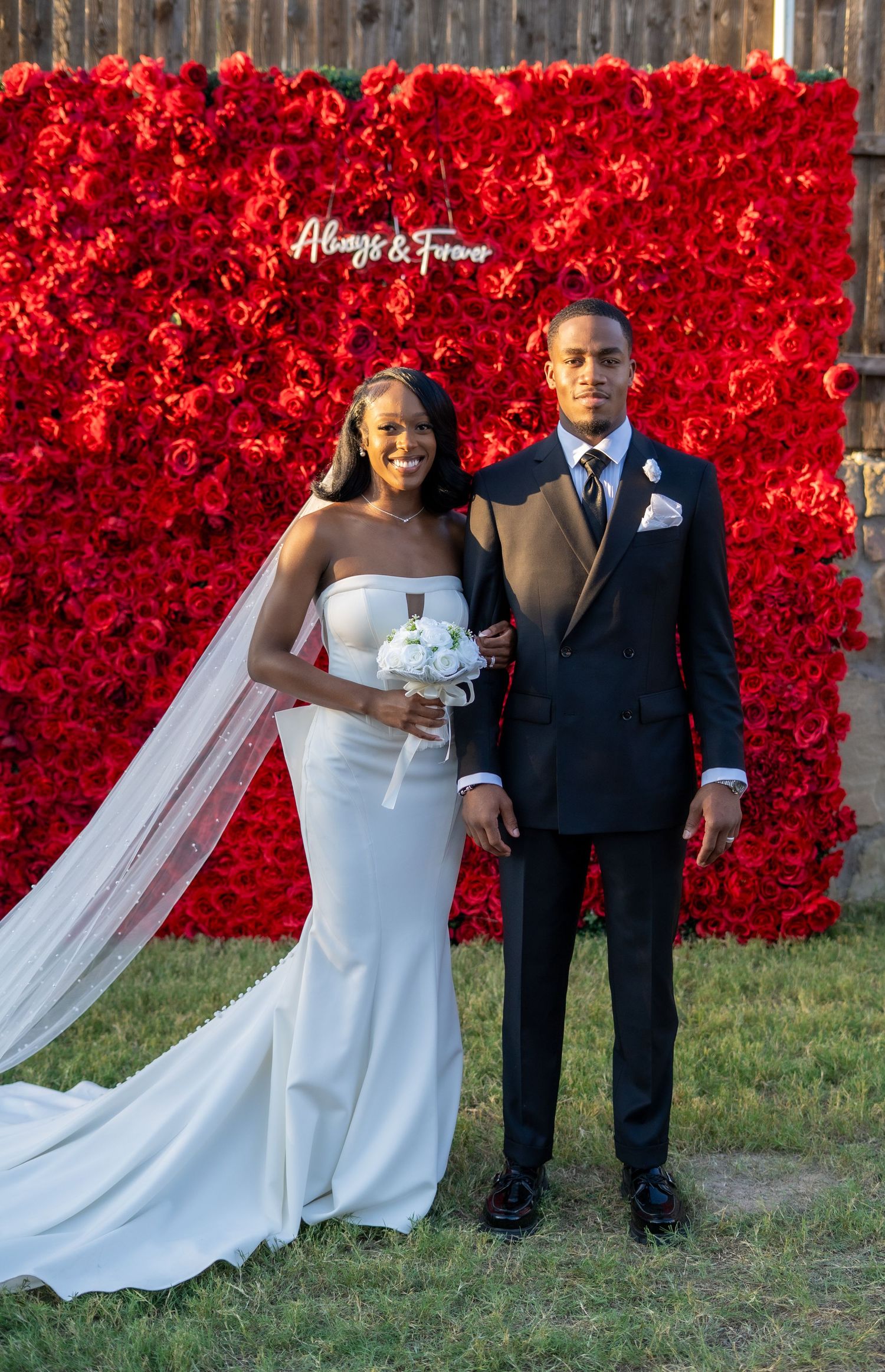 Bride holding white bouquet and groom pose together in front of red rose flower wall backdrop.