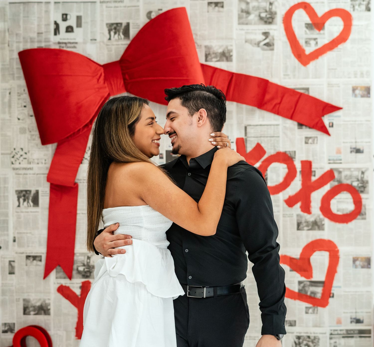 Couple embracing and smiling at each other in front of Valentine's Day backdrop with red bow.