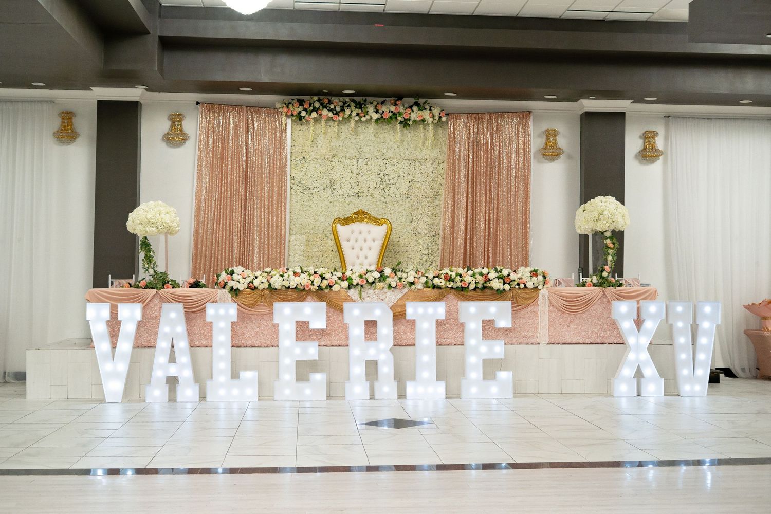Elegant Quinceañera venue with VALERIE XV marquee letters, floral backdrop, and gold throne chair.