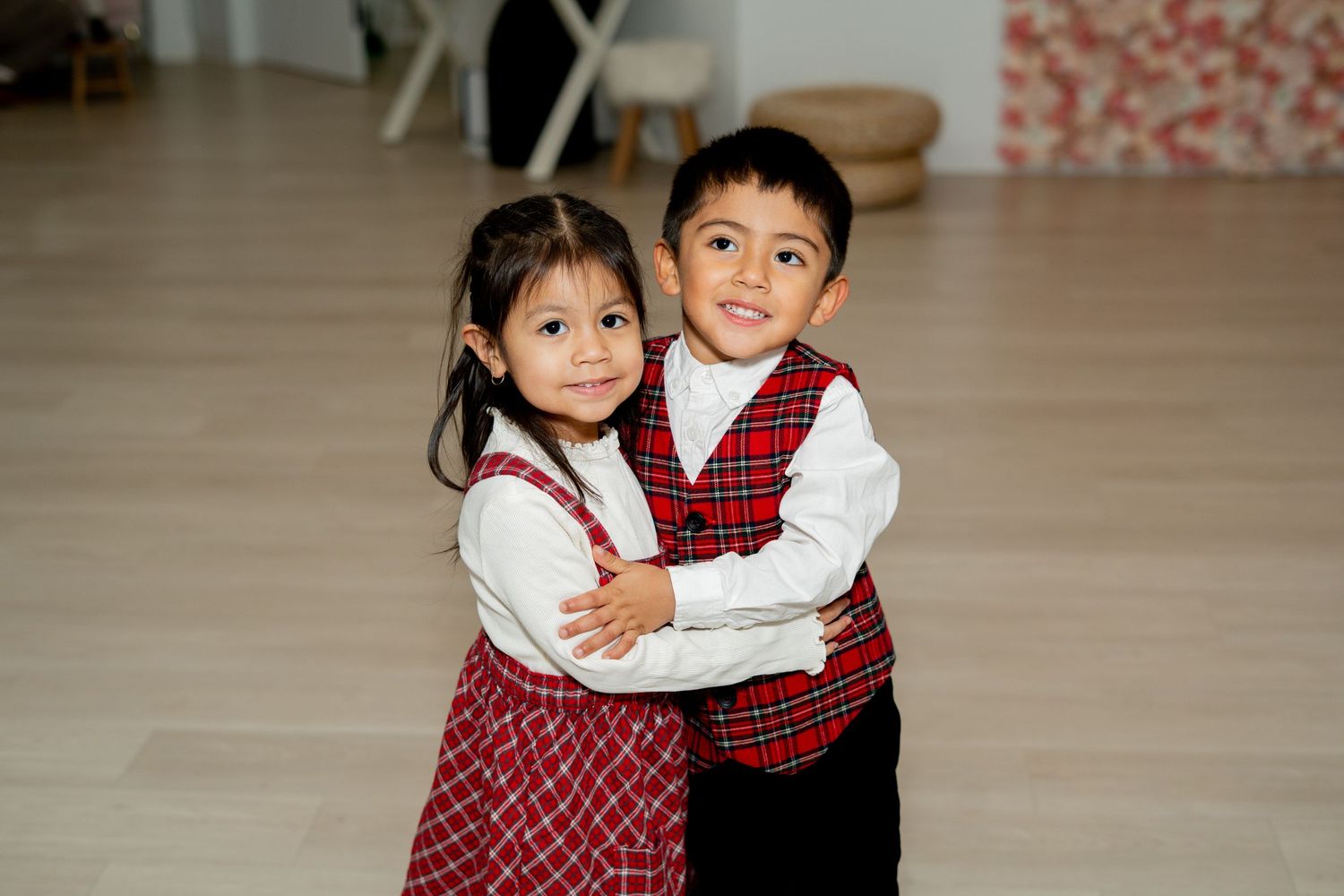 Two young children in matching red plaid holiday outfits hugging and smiling in a bright studio.