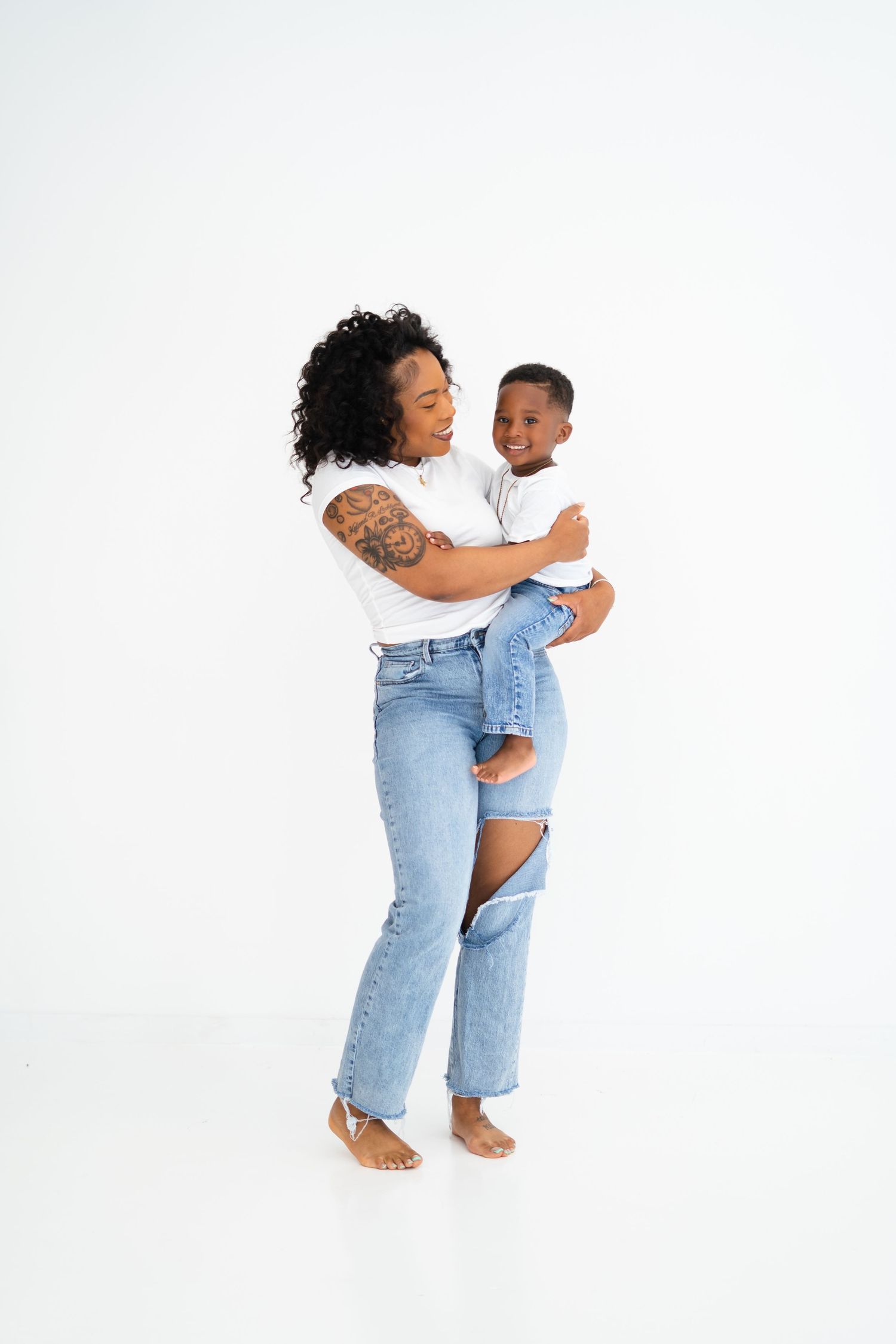 Black mother with tattoos holding her smiling young son in matching white outfits in studio.