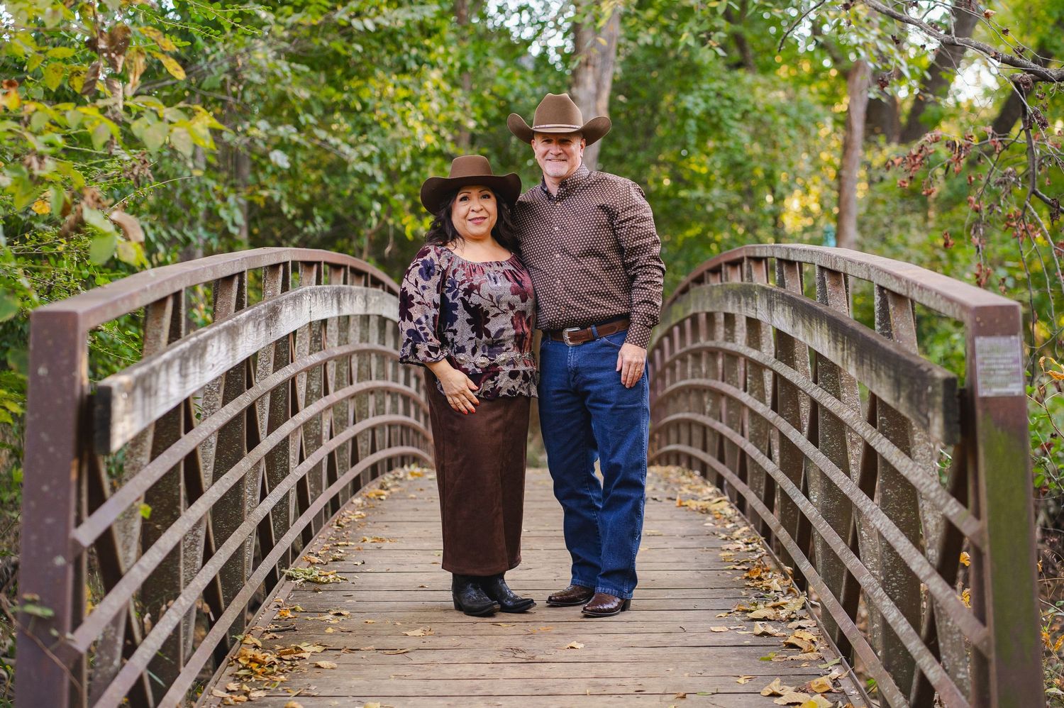 Couple wearing cowboy hats posing together on a wooden bridge surrounded by lush autumn trees.