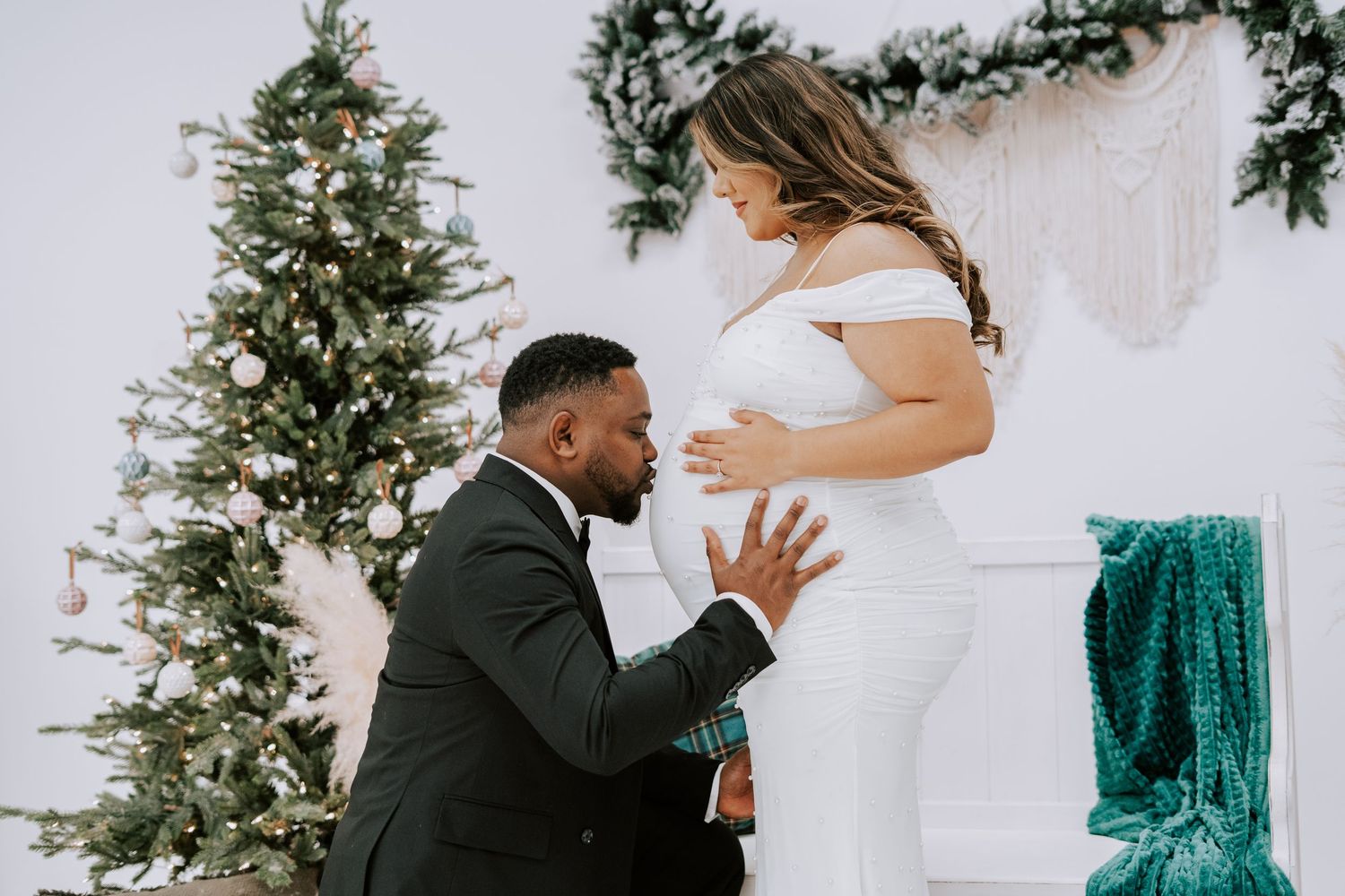 Man in tuxedo kneeling to kiss pregnant woman's belly beside Christmas tree and white fireplace mantel.