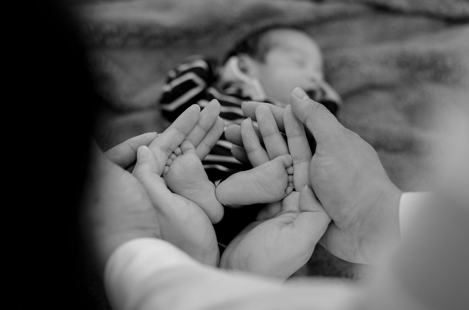 Black and white image of parents holding newborn baby's feet with sleeping infant in background.