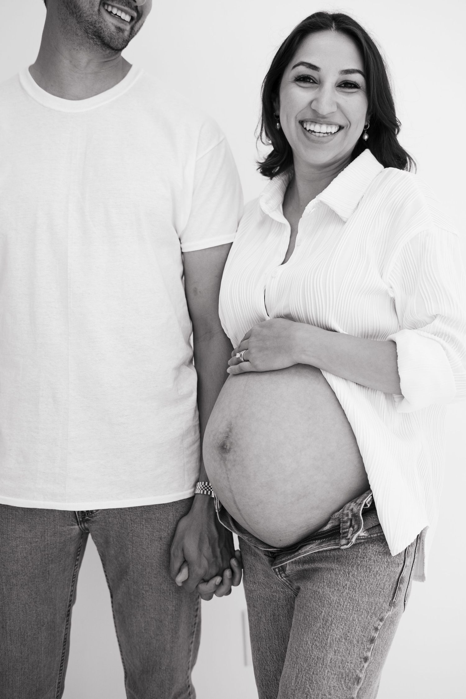 Black and white photo of a smiling pregnant woman holding hands with a man, cradling her bare baby bump.