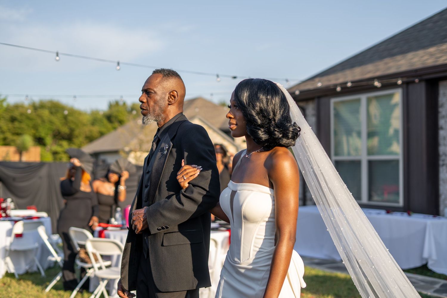 Father of the bride walks daughter in white wedding gown and veil toward outdoor ceremony venue.