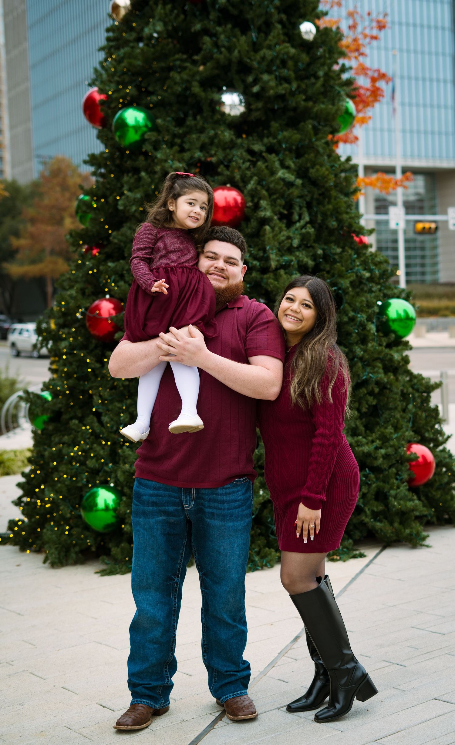 Family of three in coordinating burgundy outfits poses by a large decorated outdoor Christmas tree.