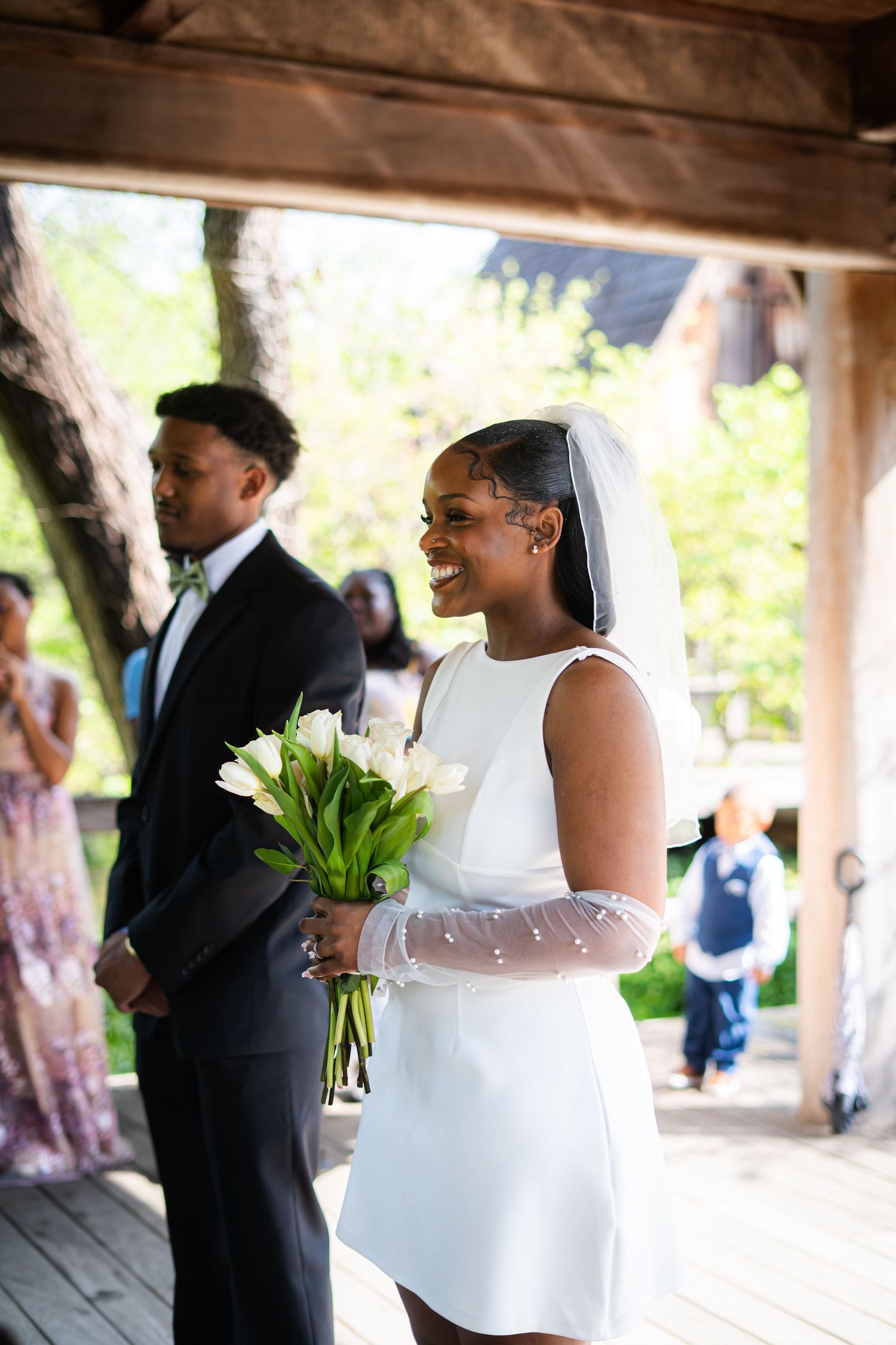 Radiant bride in short white dress and veil holding white tulips laughs joyfully during outdoor wedding ceremony.