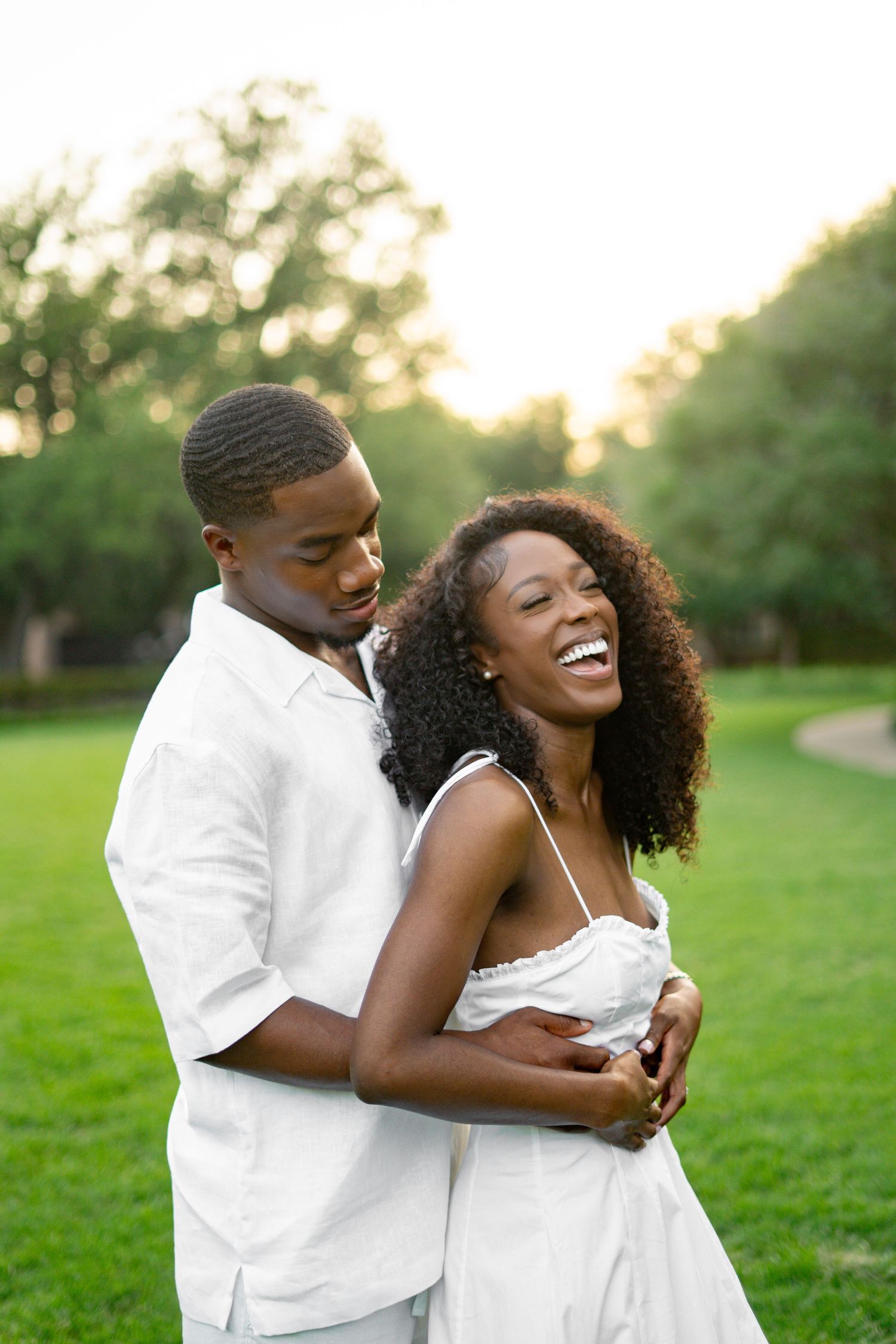 Laughing Black woman in white dress embraced from behind by man in white shirt on green lawn.