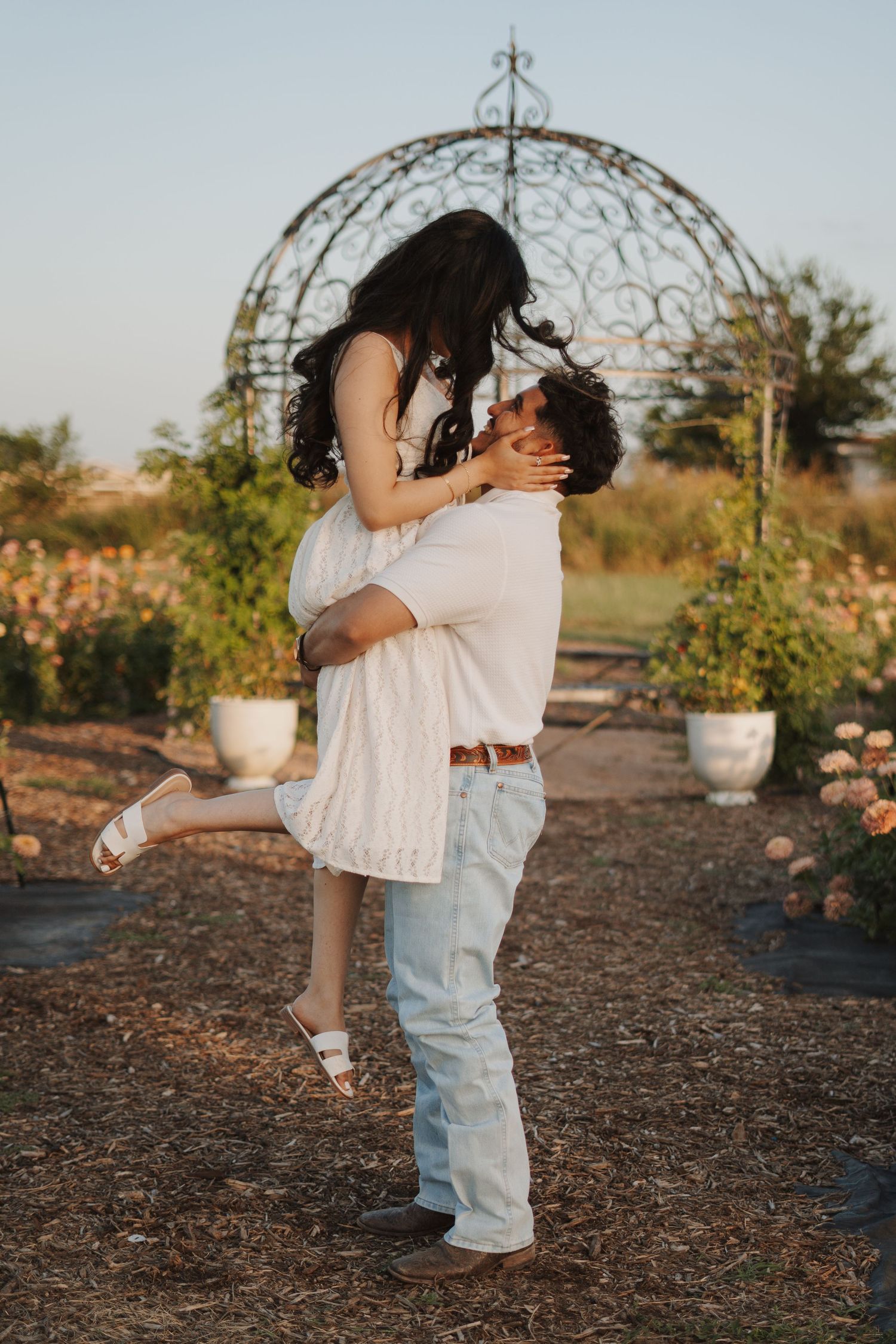 A couple shares a romantic moment as a man lifts a woman in a white lace dress in a garden.
