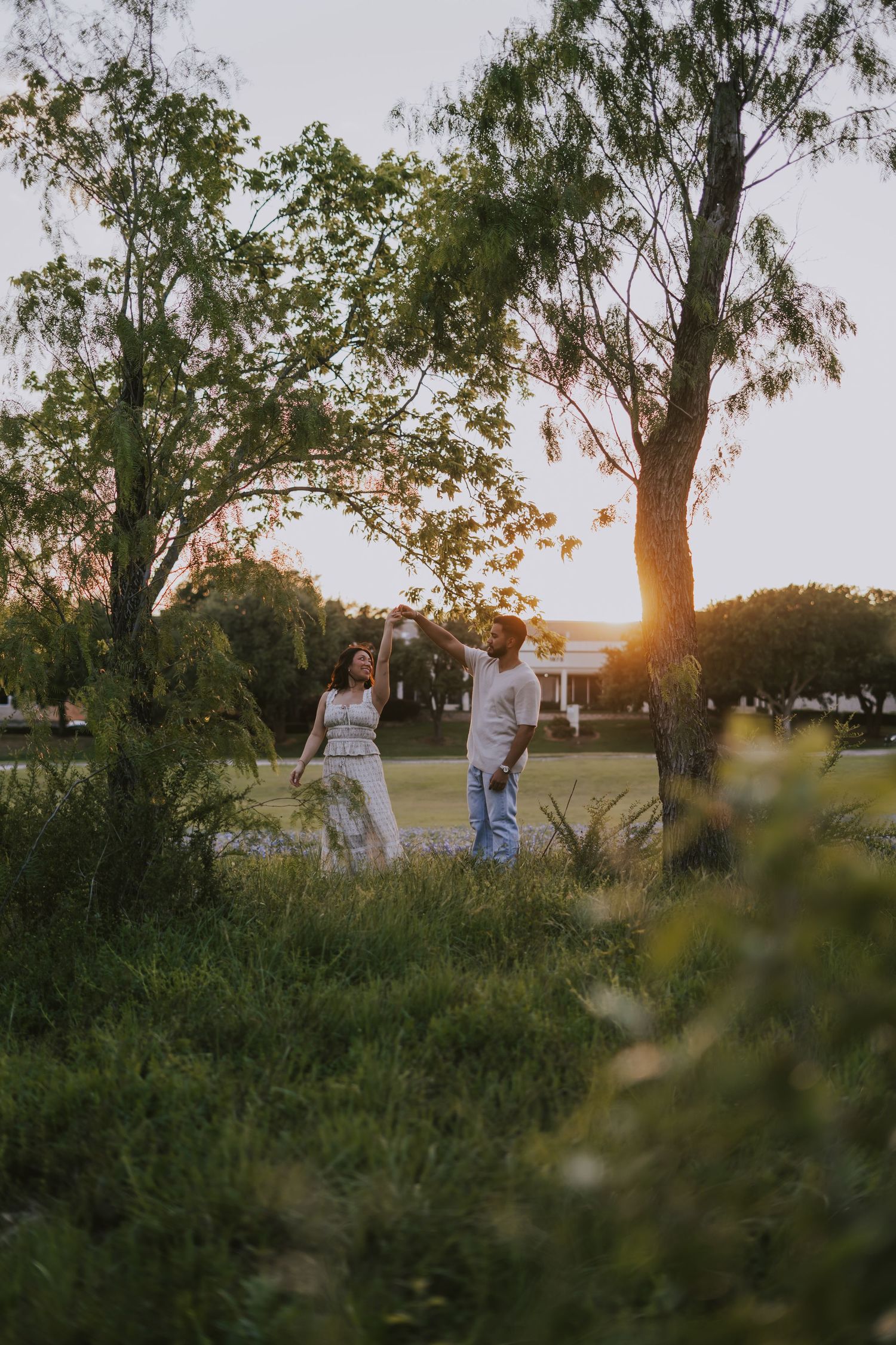 Couple dancing together in a lush green park at golden sunset, surrounded by tall trees and warm glowing light.