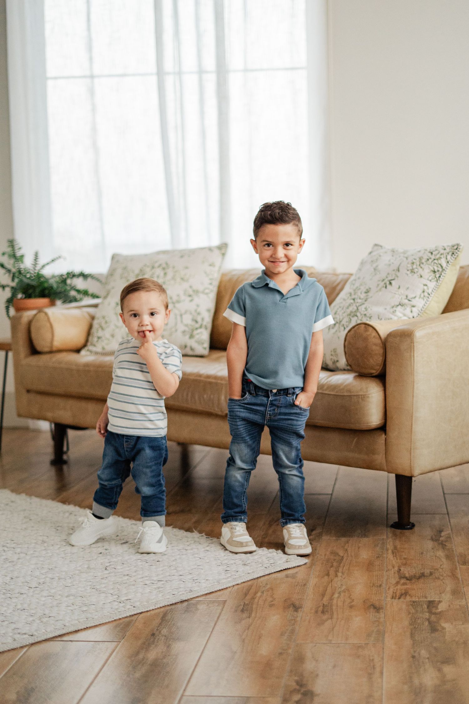 Two young boys in jeans and casual tops smile while standing in a bright living room with hardwood floors and a beige sofa.