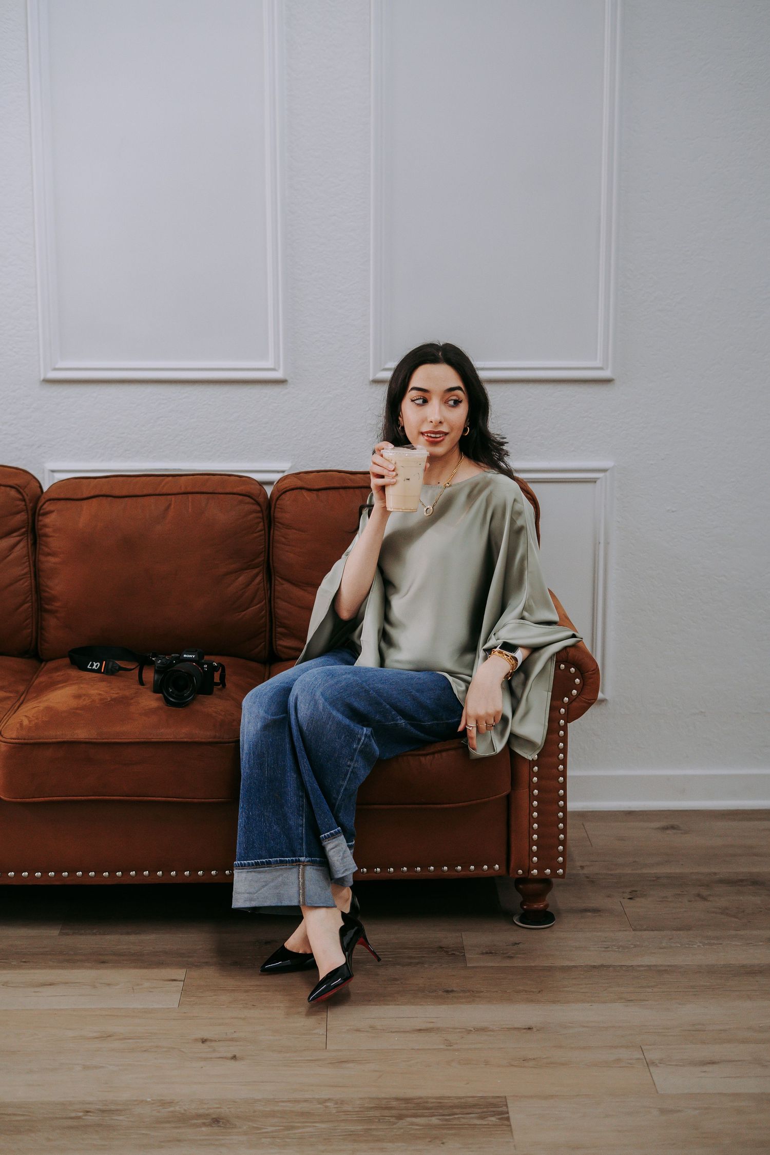 Woman in sage blouse and jeans sitting on brown leather sofa, holding a drink in a bright modern room.