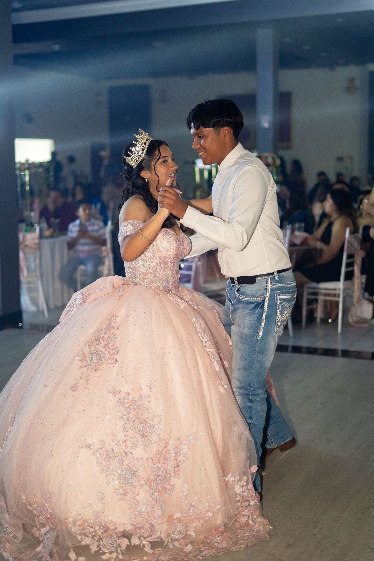 Quinceañera girl in pink floral ball gown and tiara shares romantic waltz with chambelan at party.