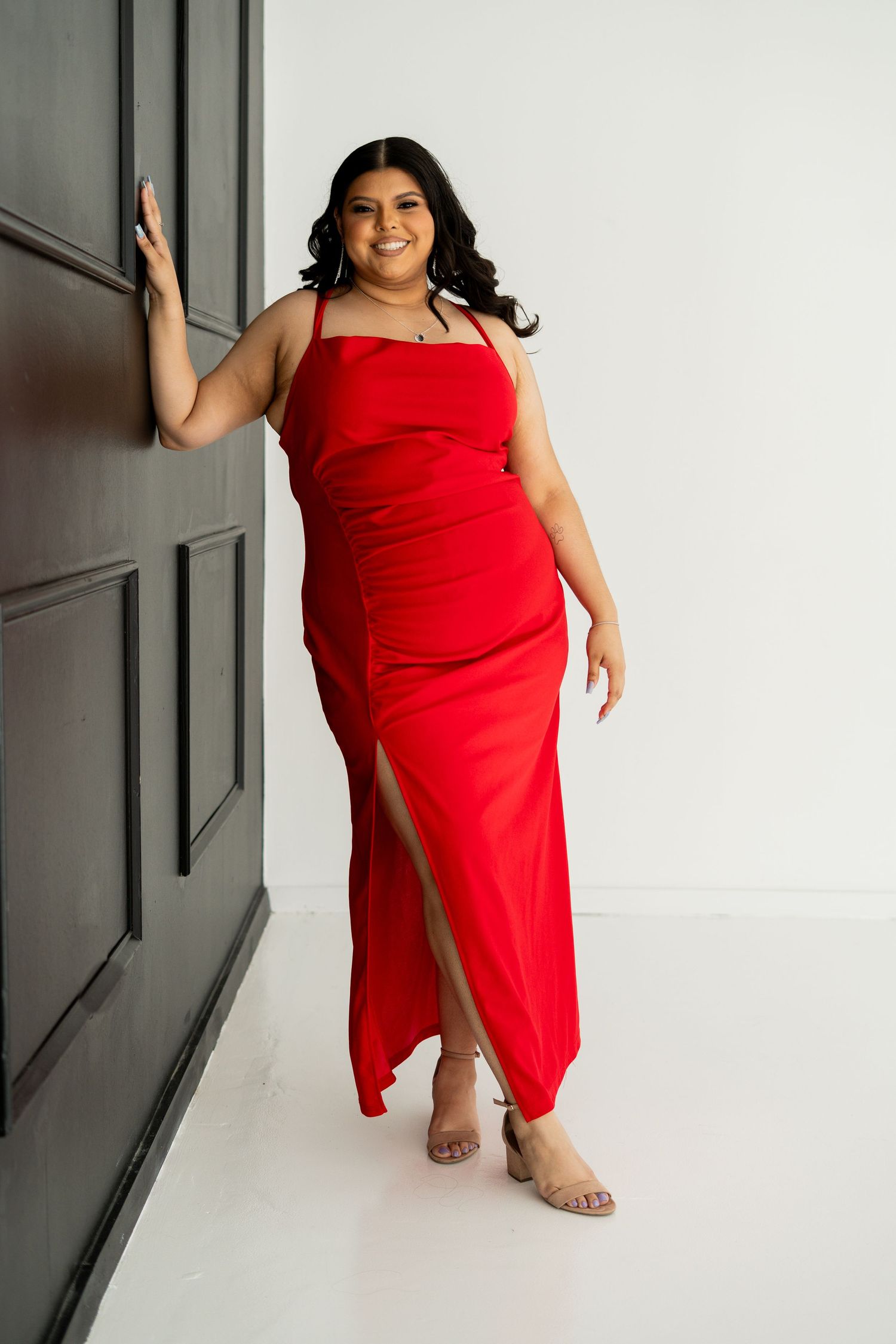 Woman in a red sleeveless slit maxi dress posing confidently beside a dark door in a studio setting.