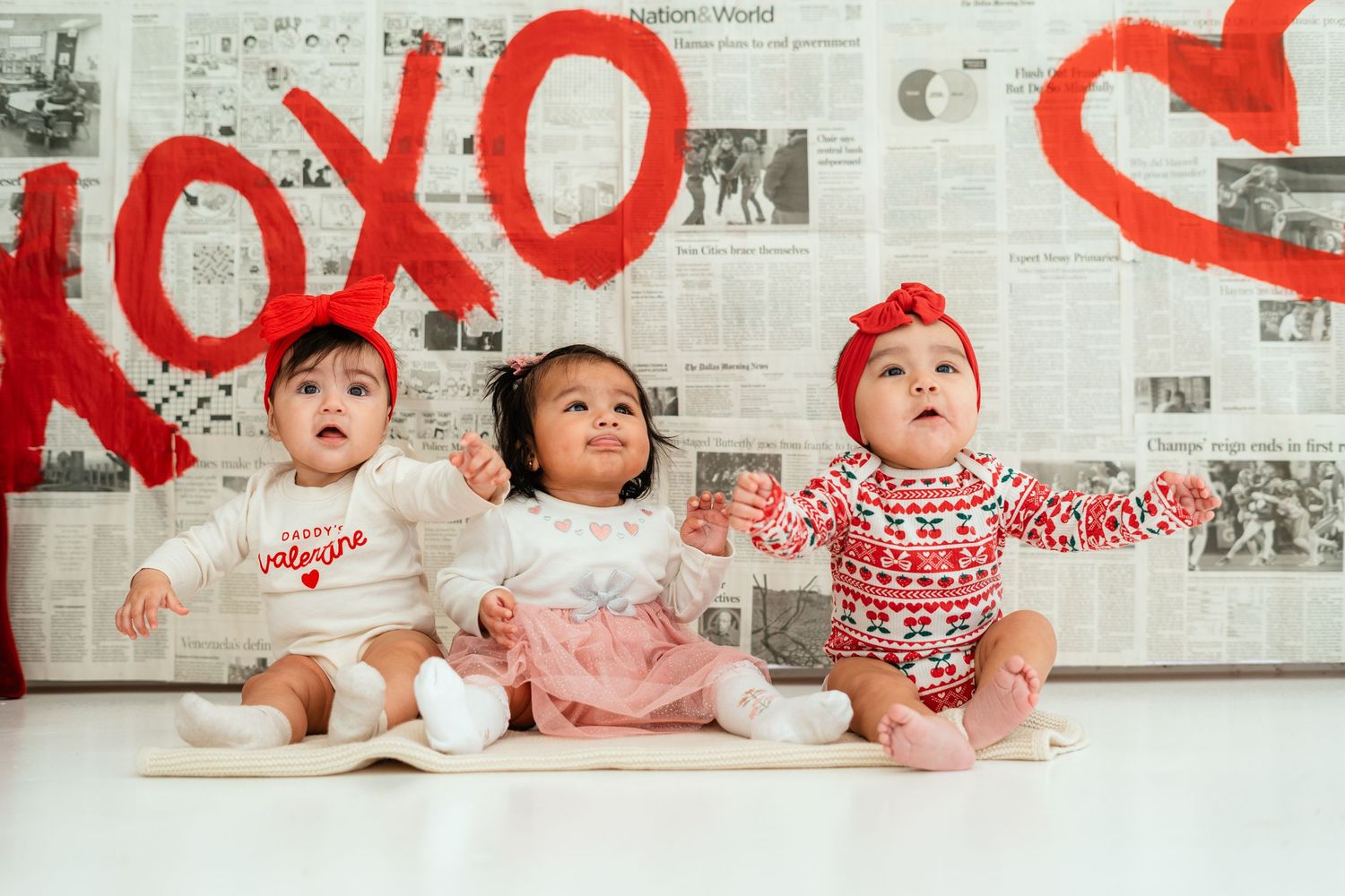 Three adorable babies in Valentine's Day outfits sitting before a red XOXO backdrop.