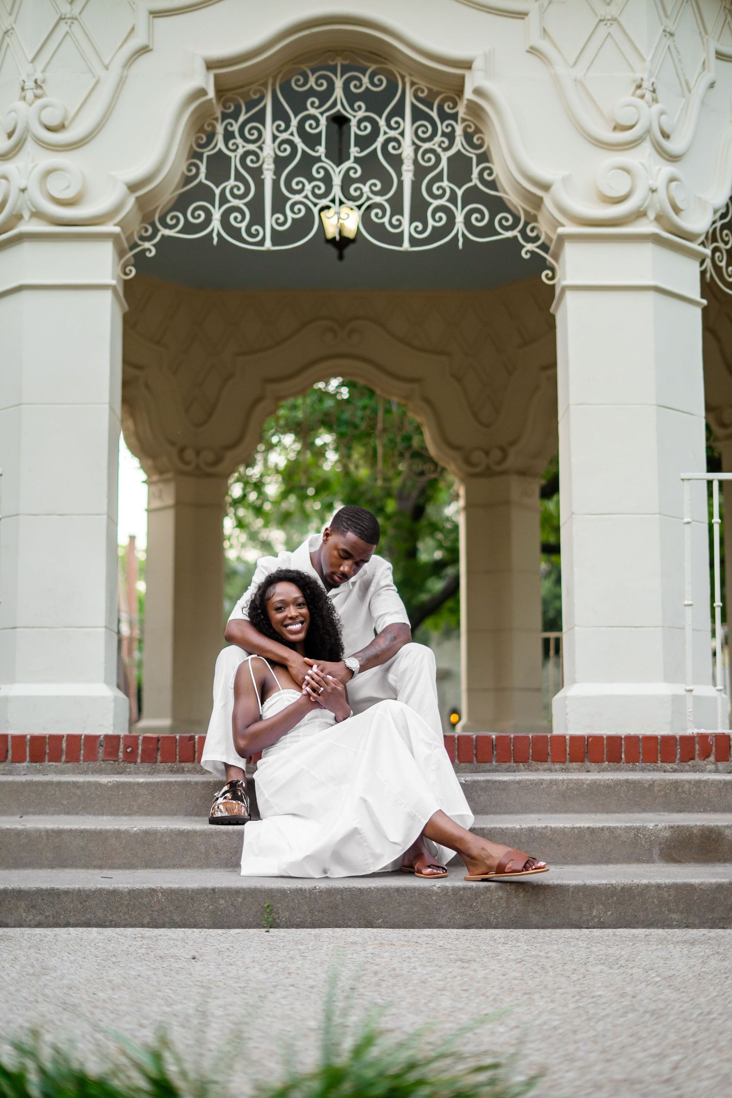 A smiling Black couple in white outfits pose together on steps beneath an elegant white decorative gazebo.
