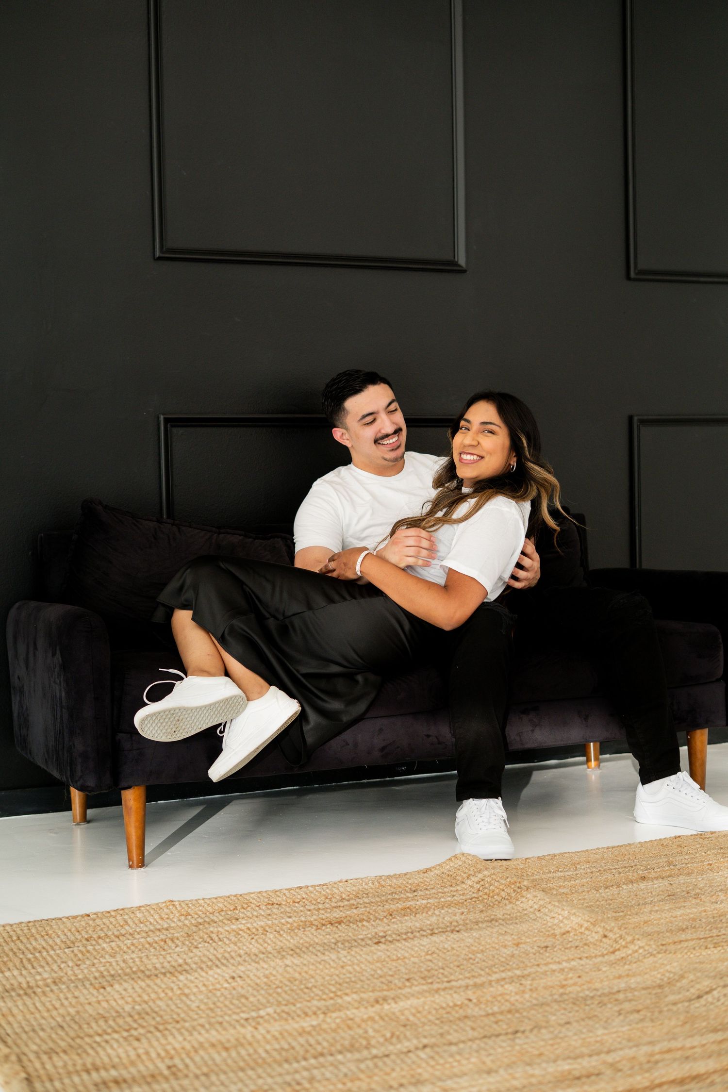 Joyful couple cuddling on a black velvet sofa against a dark paneled wall.