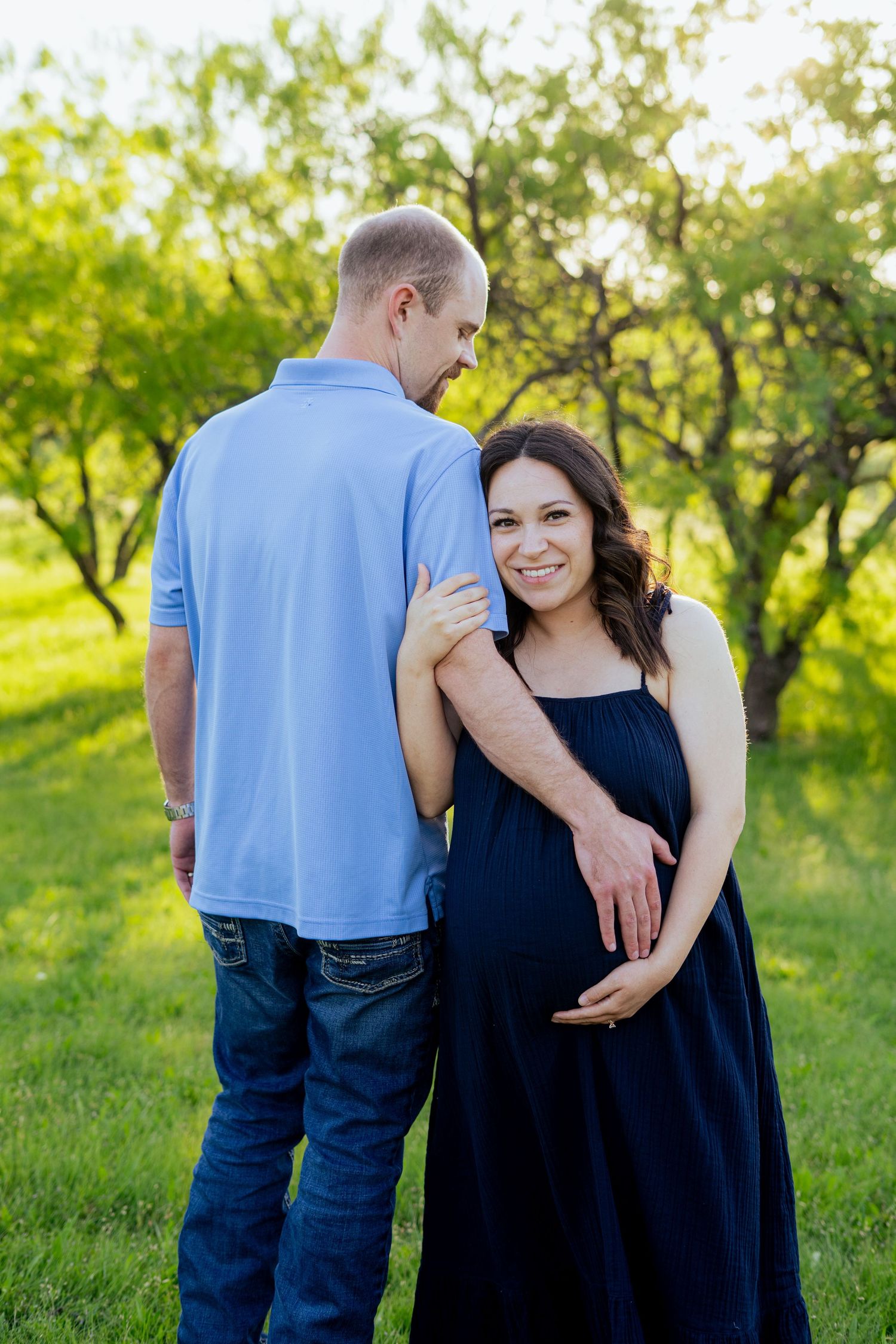 Smiling pregnant woman in navy dress posing with her partner in a lush green park.