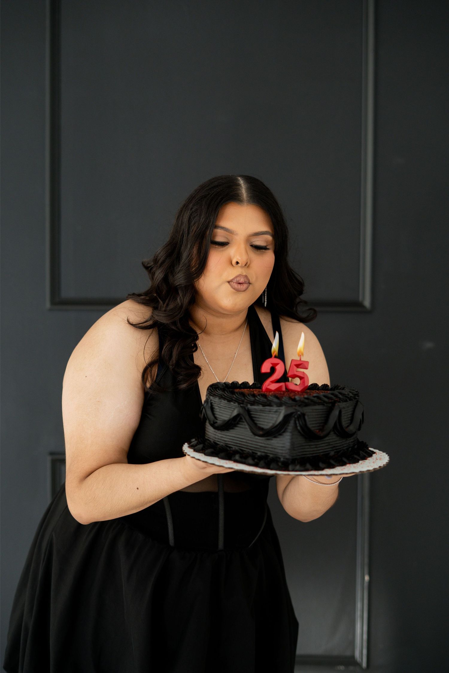 Woman in black dress blowing out candles on a black birthday cake with number 25 candles.